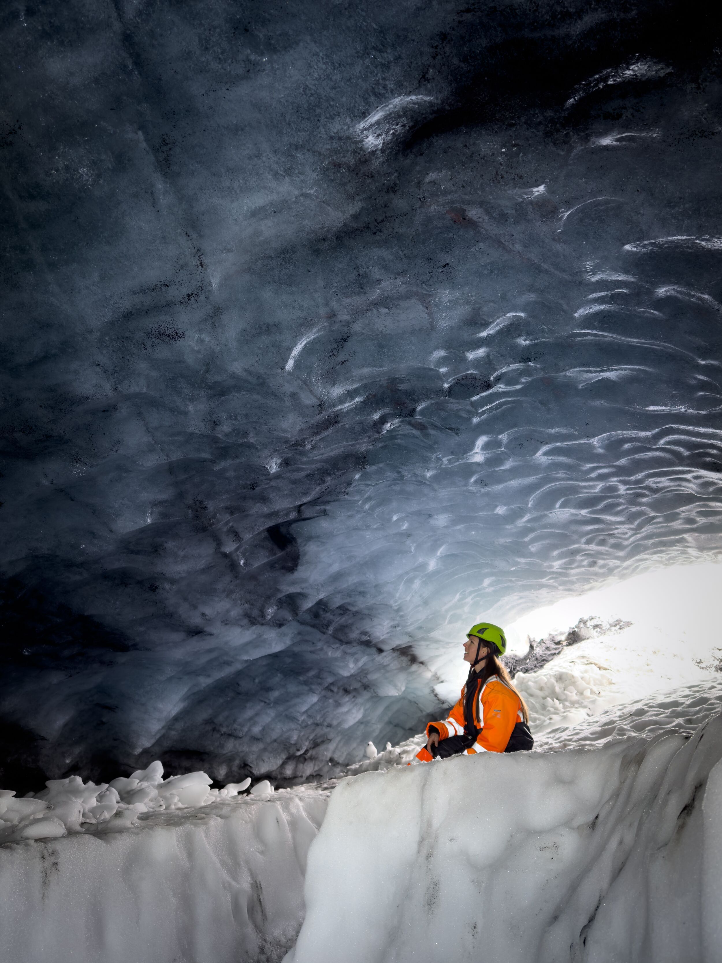 Person in orange and black suit and green helmet exploring Askur ice cave surrounded by ice and snow.