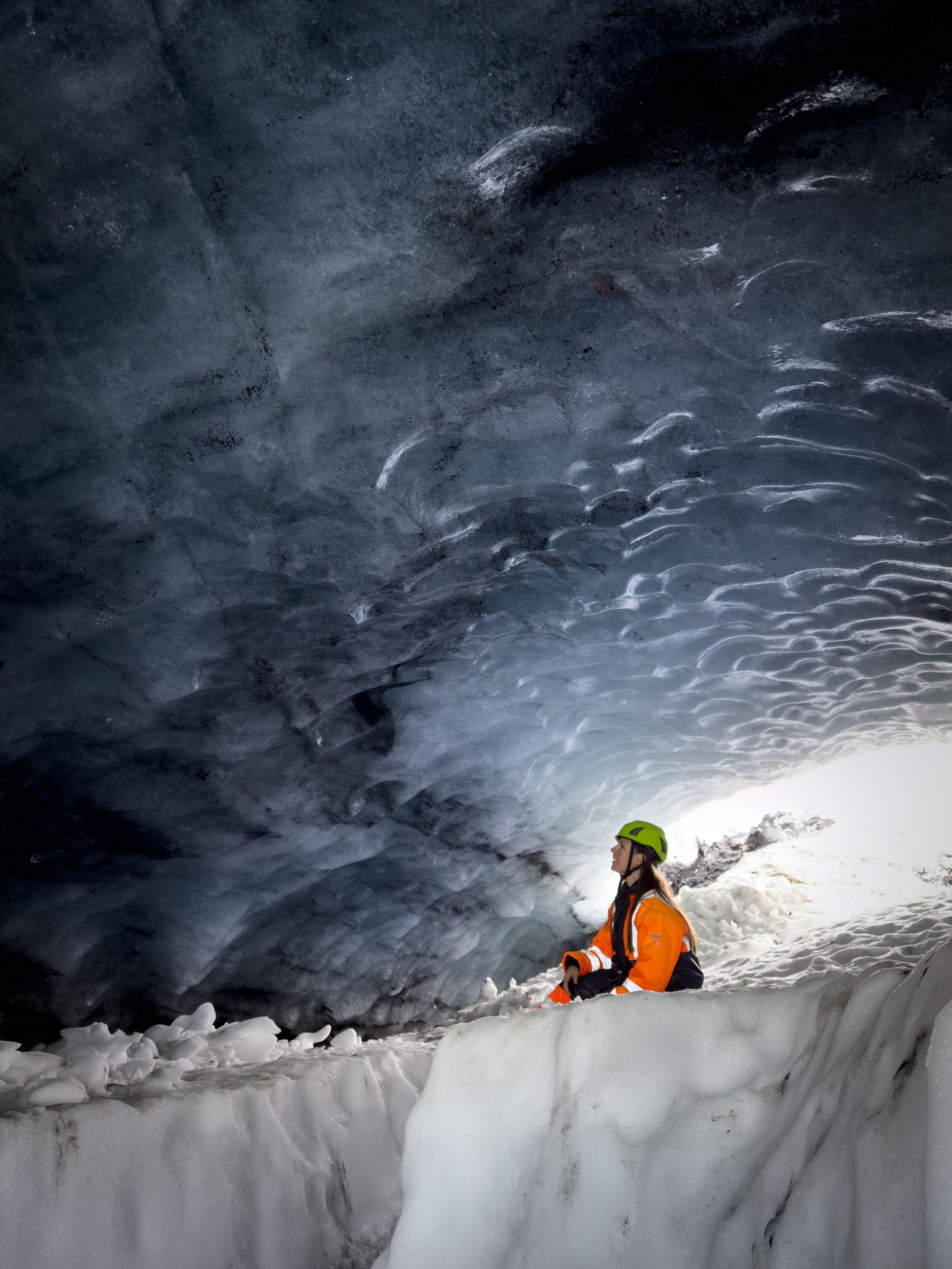 Person in a green hat and oange jacket sitting inside Askur ice cave, surrounded by stunning blue ice formations.