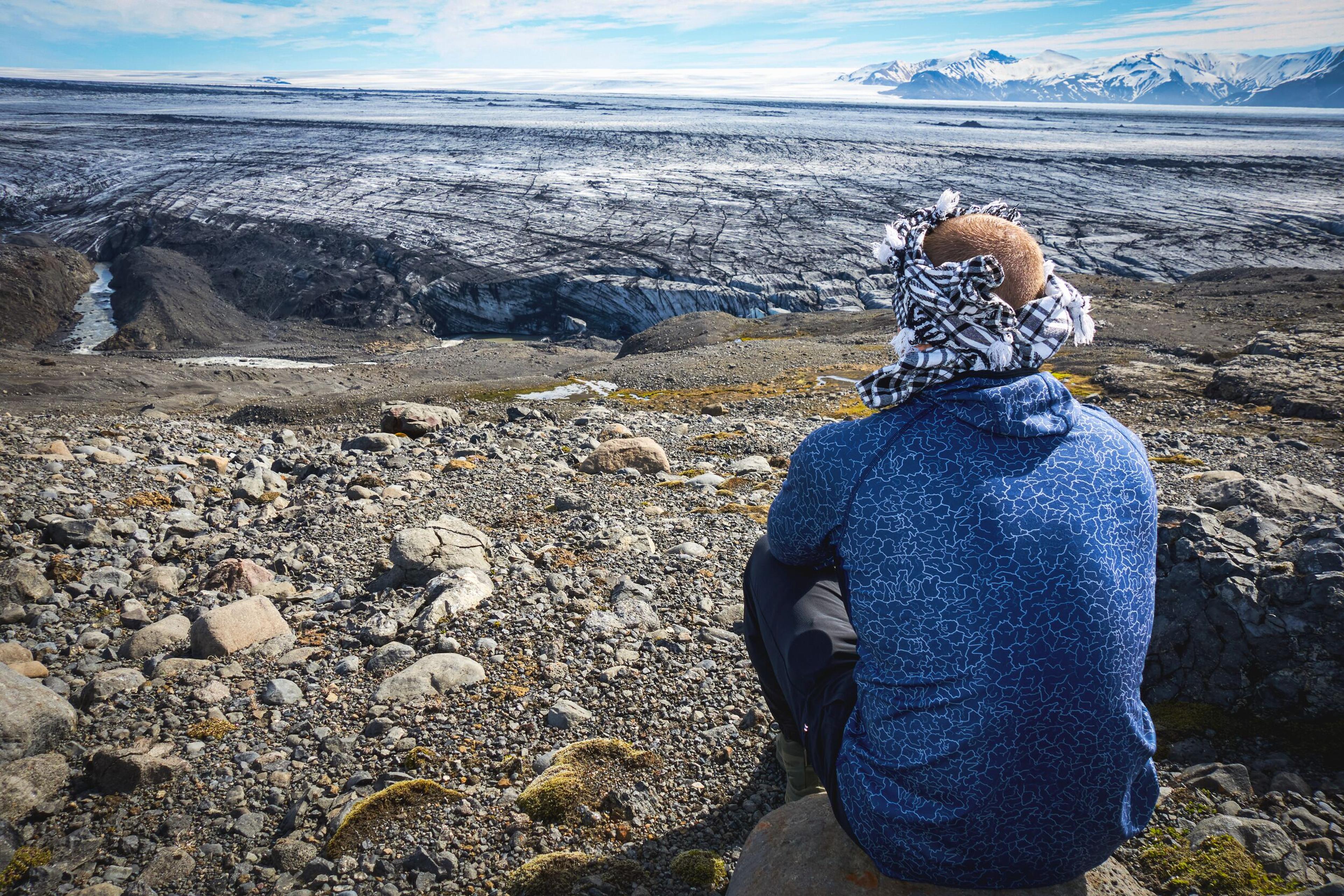 A person from behind, wearing a patterned head covering, sits on a rock overlooking a vast, dark glacier and distant snow-capped mountains.