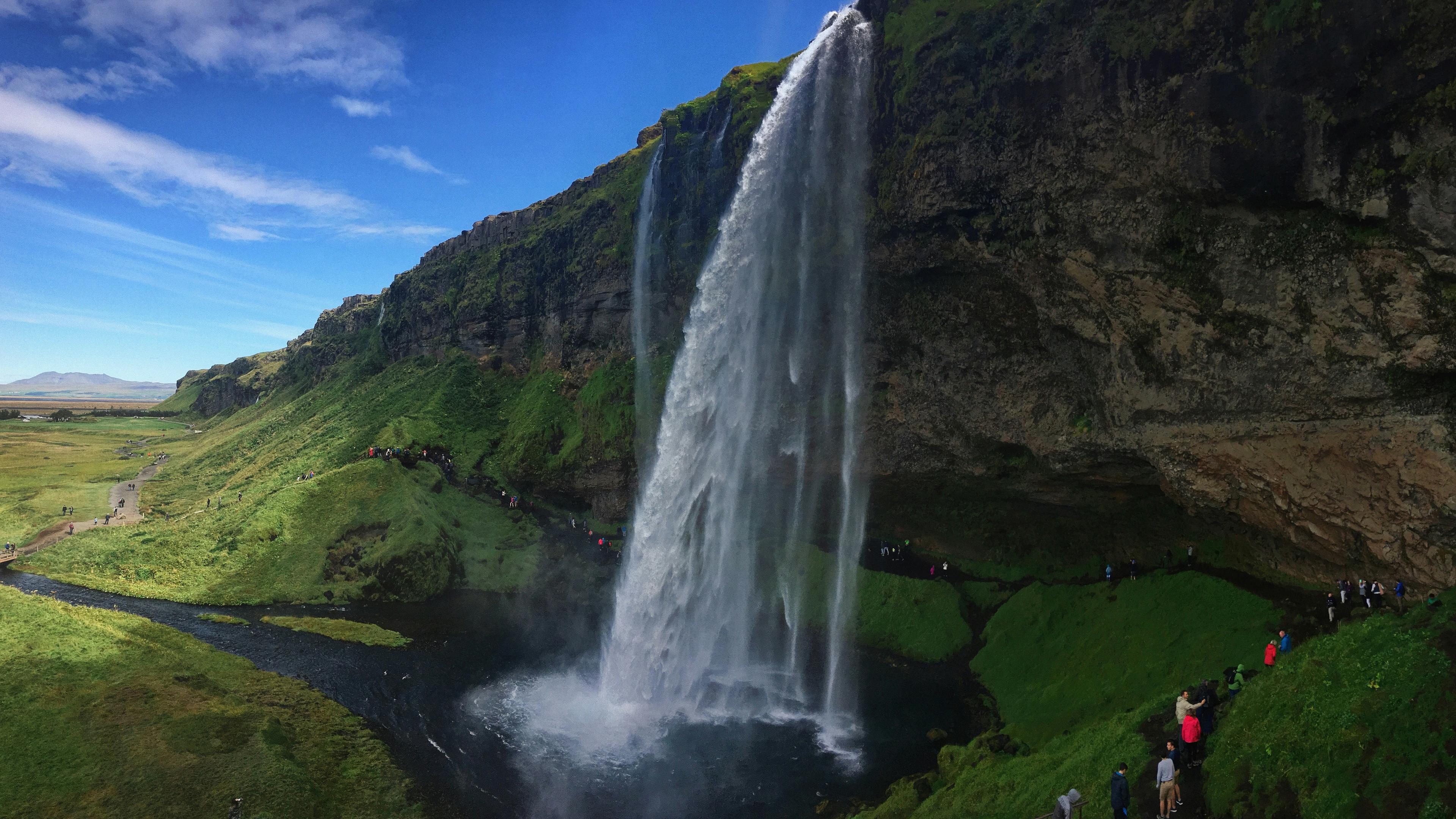 A side view of Seljalandsfoss waterfall in Iceland, showing its powerful cascade over a moss-covered cliff with a walking path behind the falls and visitors exploring the area.