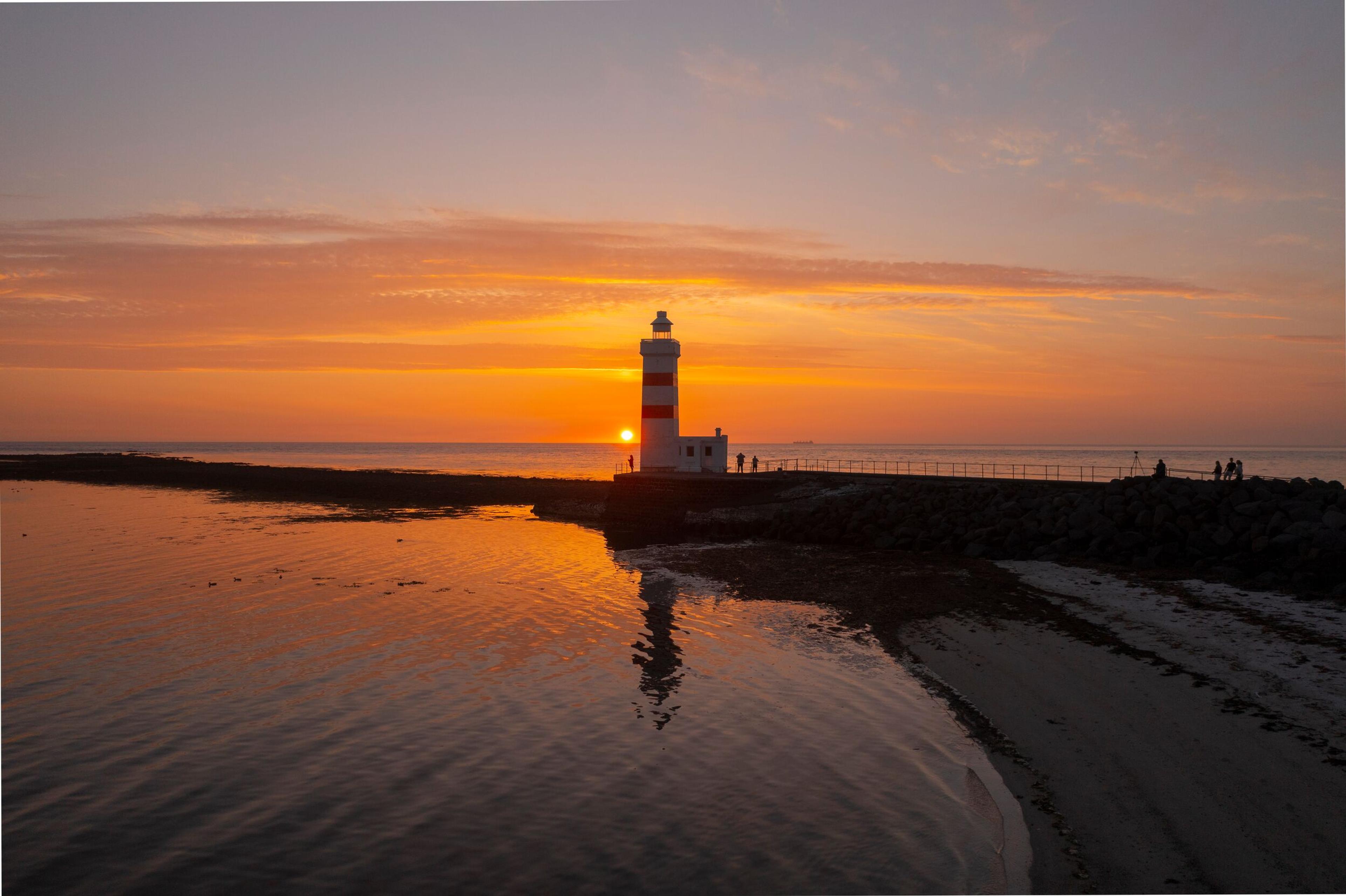 The Garður lighthouse in Iceland stands against a stunning sunset, reflecting golden hues on the calm water and casting a peaceful coastal scene.