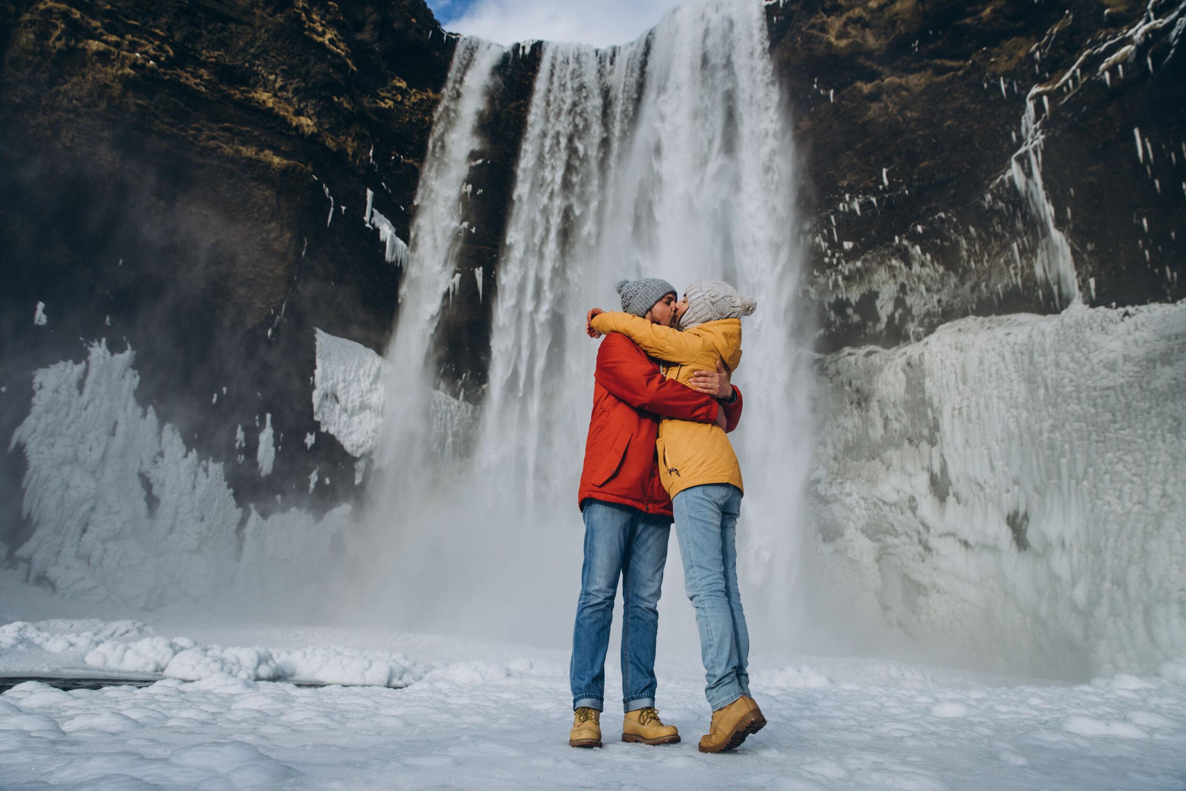 A couple in warm clothing embraces in front of Skógafoss waterfall, surrounded by ice and mist on Iceland's South Coast.