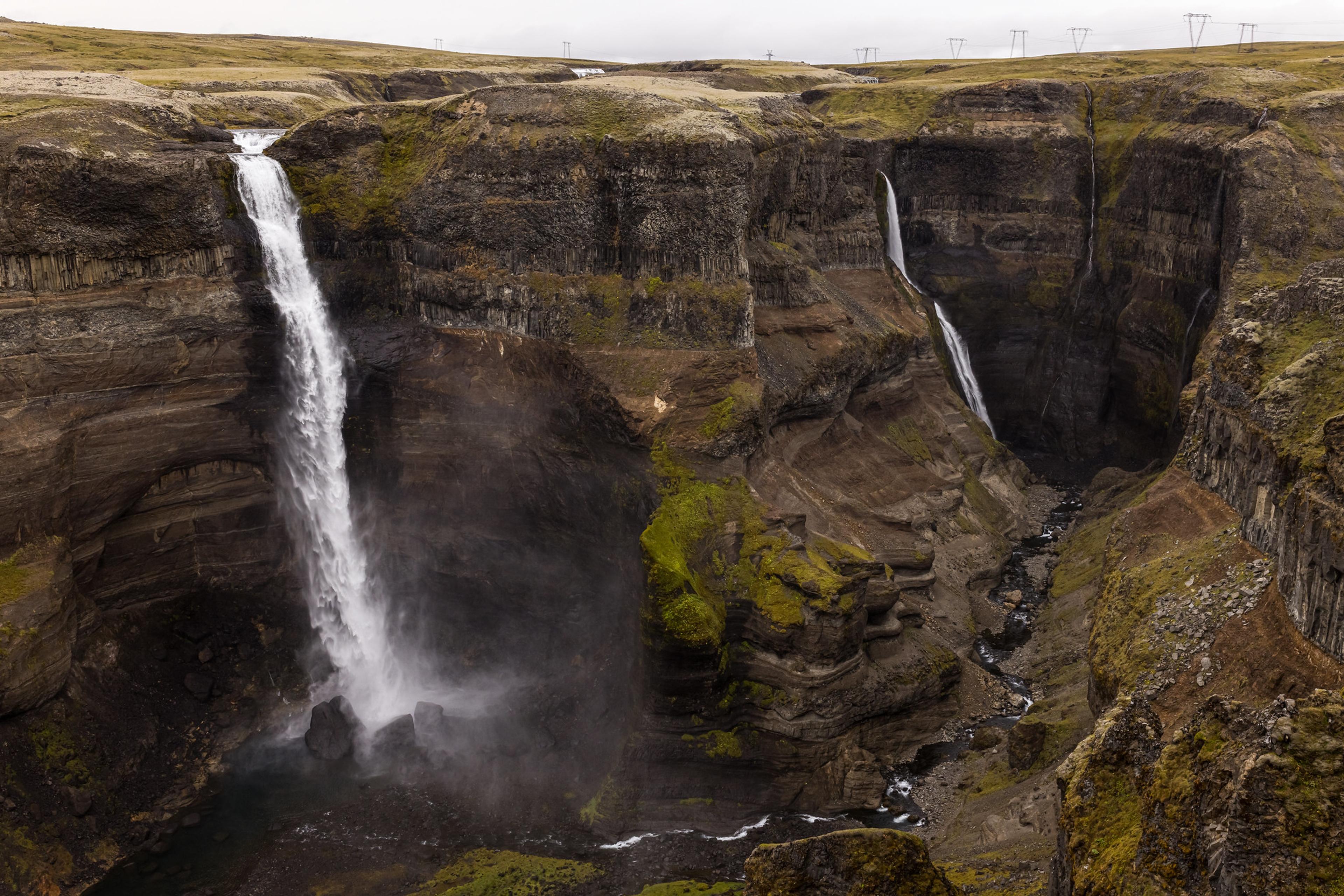 A wide waterfall and a narrower one plunge into a mossy, rocky canyon under a gray sky.