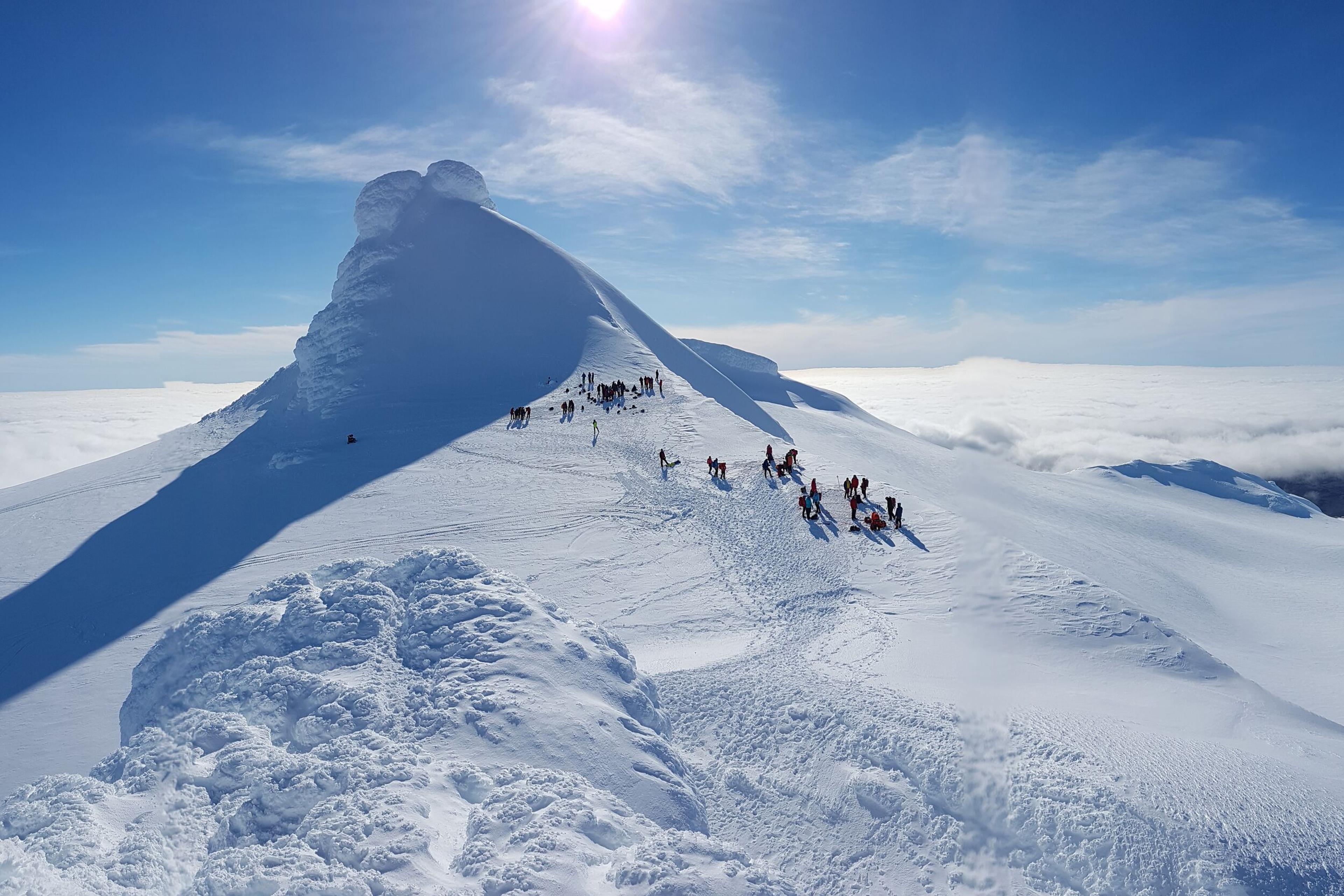  A line of climbers makes their way up a narrow snowy ridge, under a bright sun with a clear blue sky, emphasizing the vastness and isolation of the icy landscape.