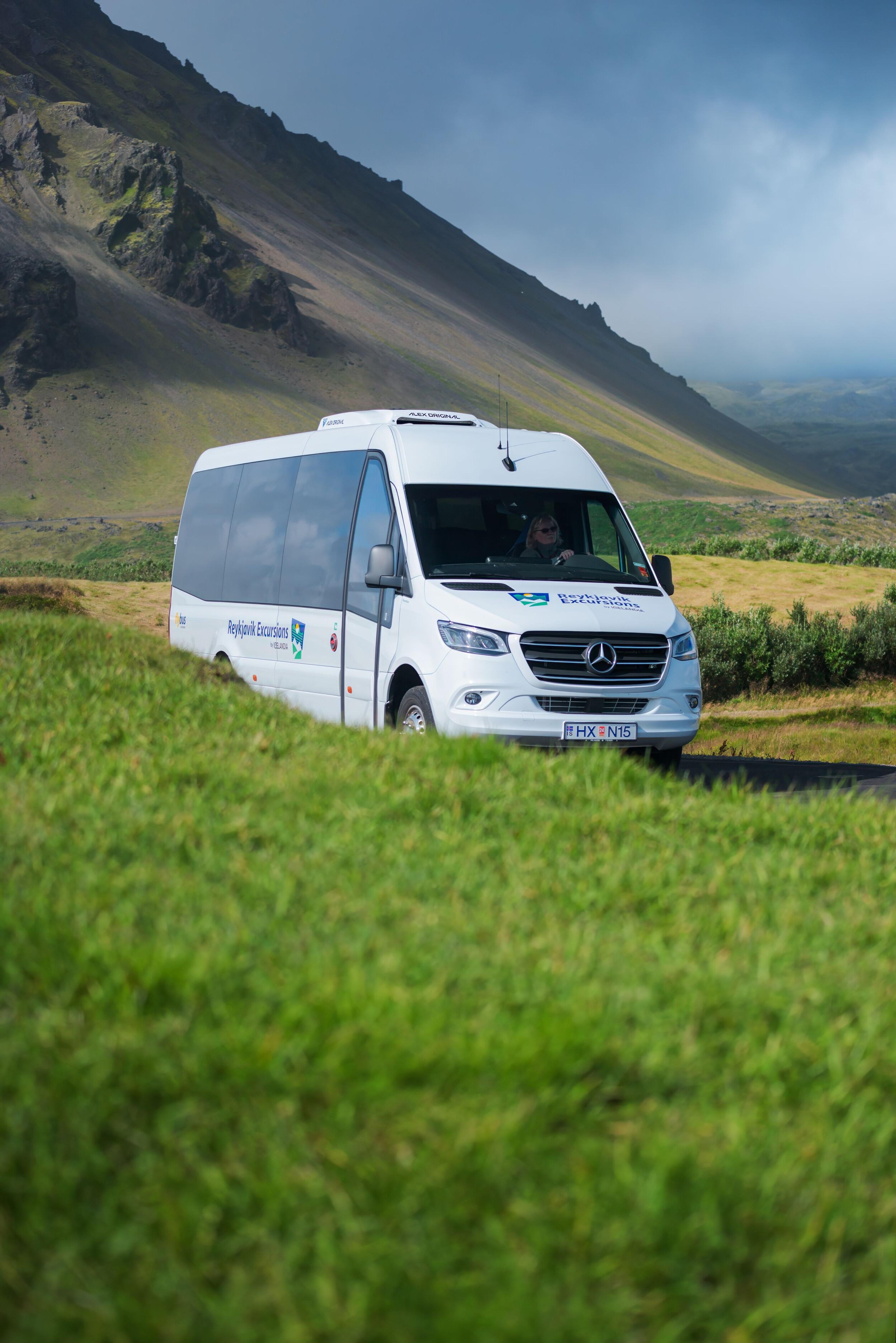 A white Reykjavik Excursions tour bus, marked as carbon neutral, travels through a lush green landscape on a sunny day.