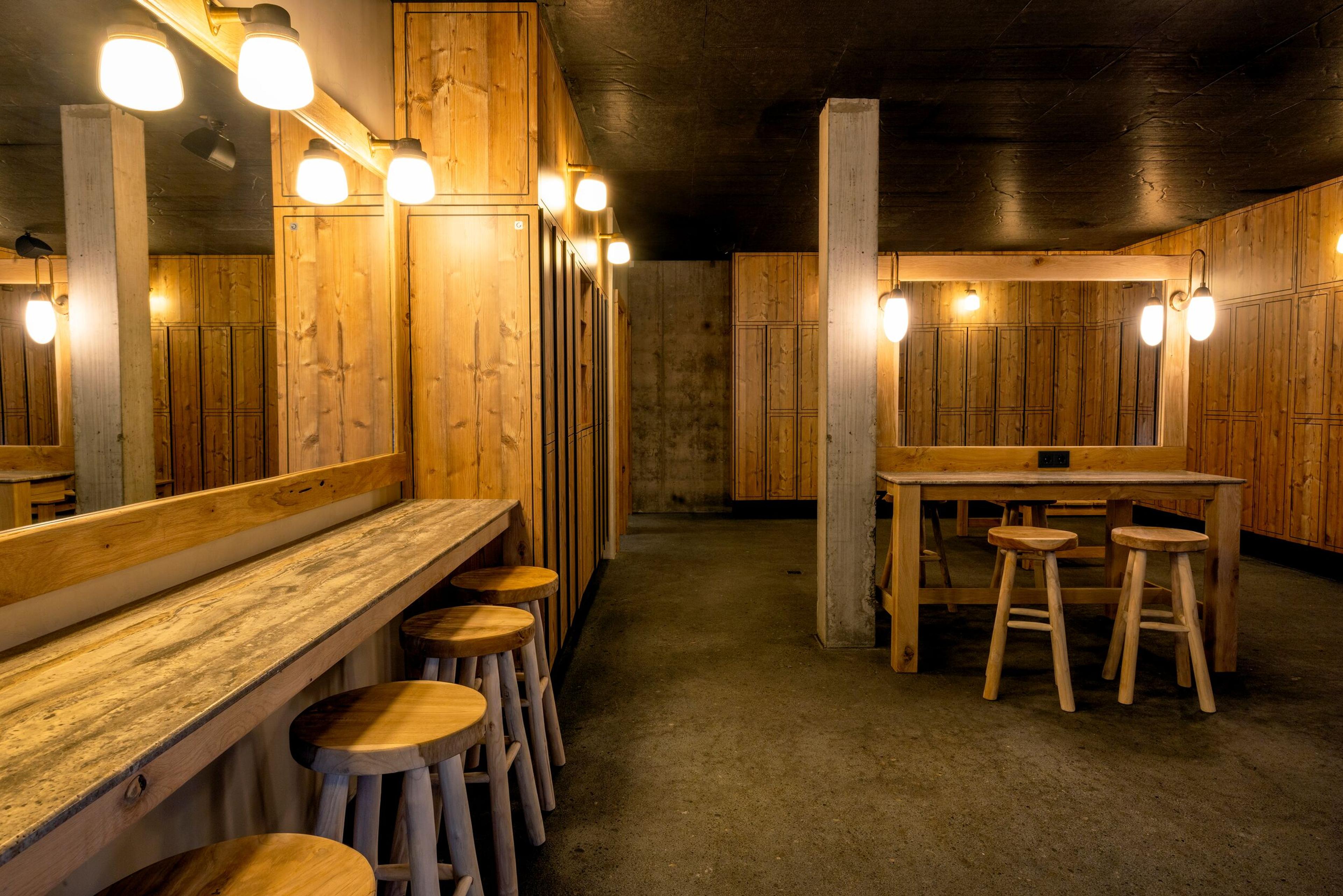 Rustic room with wooden walls, a dark ceiling, and a concrete floor, featuring a mirrored vanity counter with stools and a separate wooden table with stools.