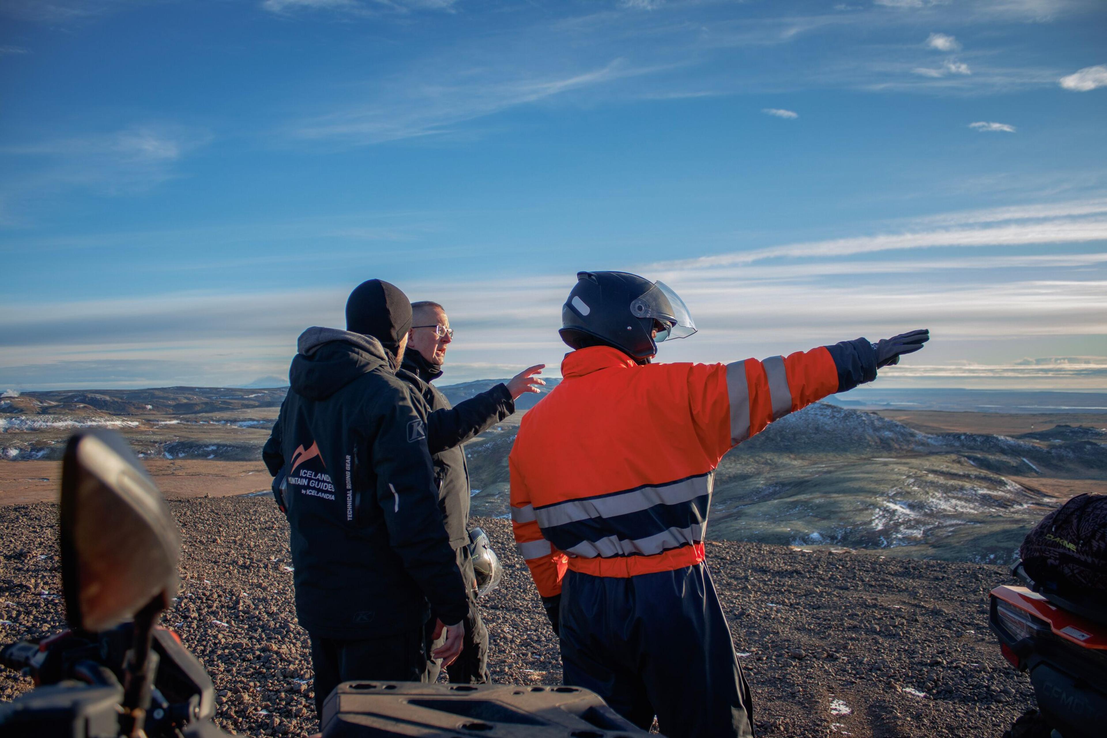 Three people on a rocky hill, one in an orange jacket and helmet points to a panoramic landscape under a blue sky.