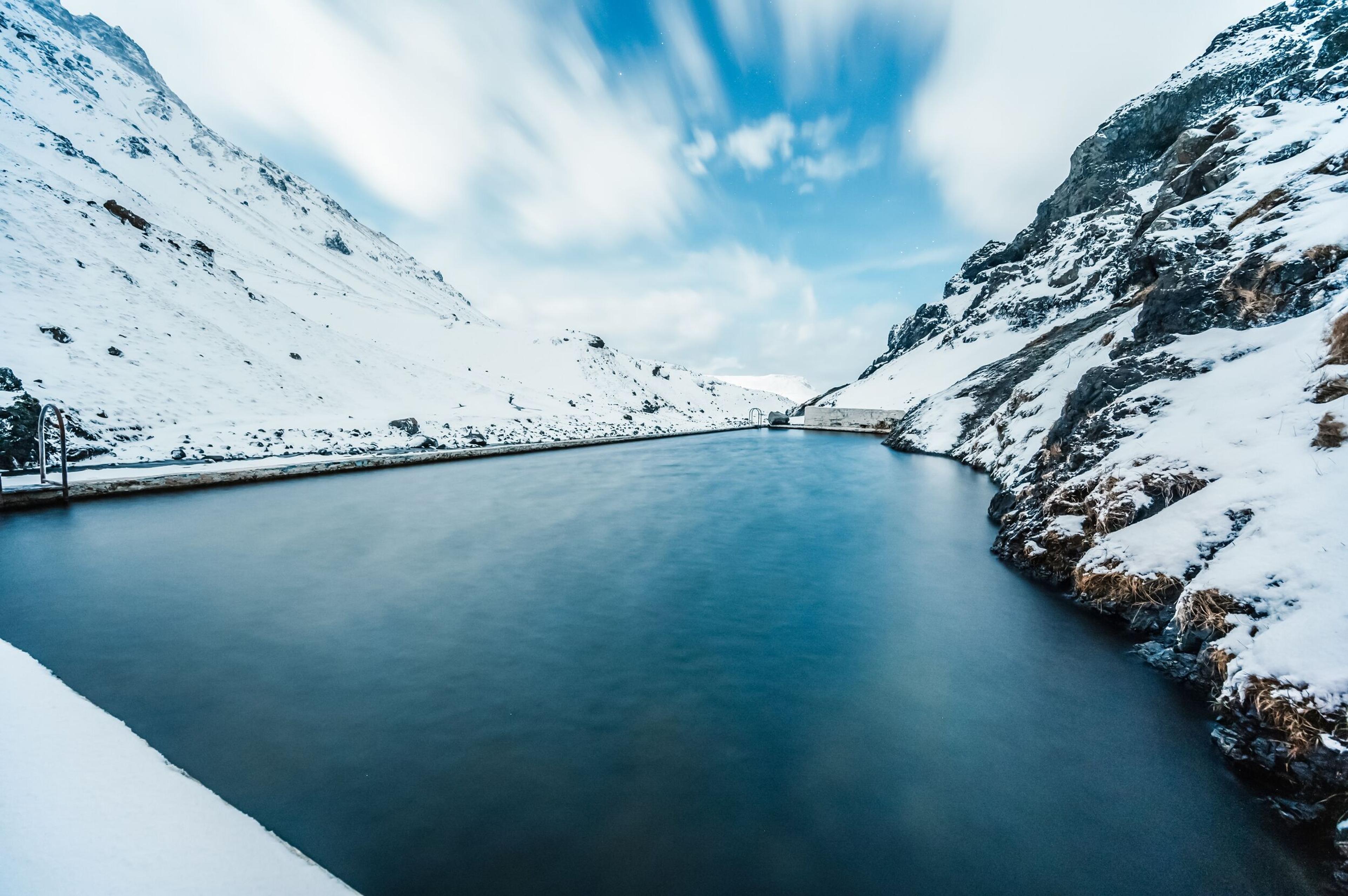 Long-exposure image of a dark lake surrounded by snow-covered mountains under a streaky sky.