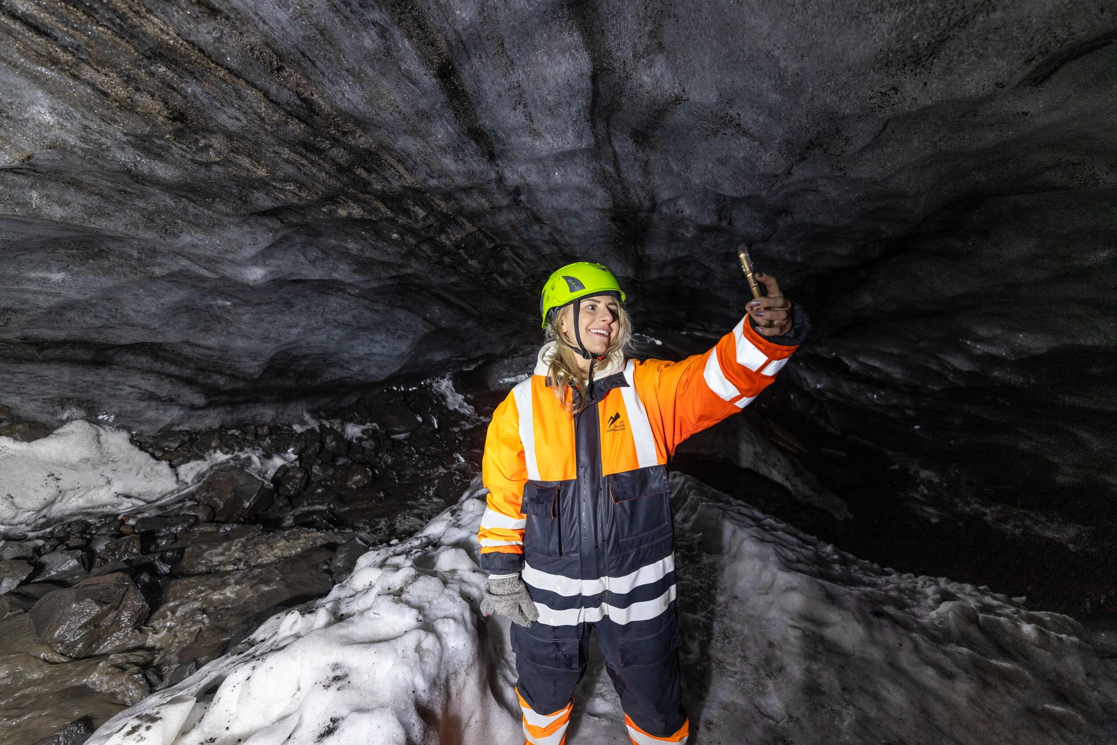 Person in orange and black suit and green helmet exploring Askur ice cave, smiling and holding a flashlight, surrounded by ice and snow.