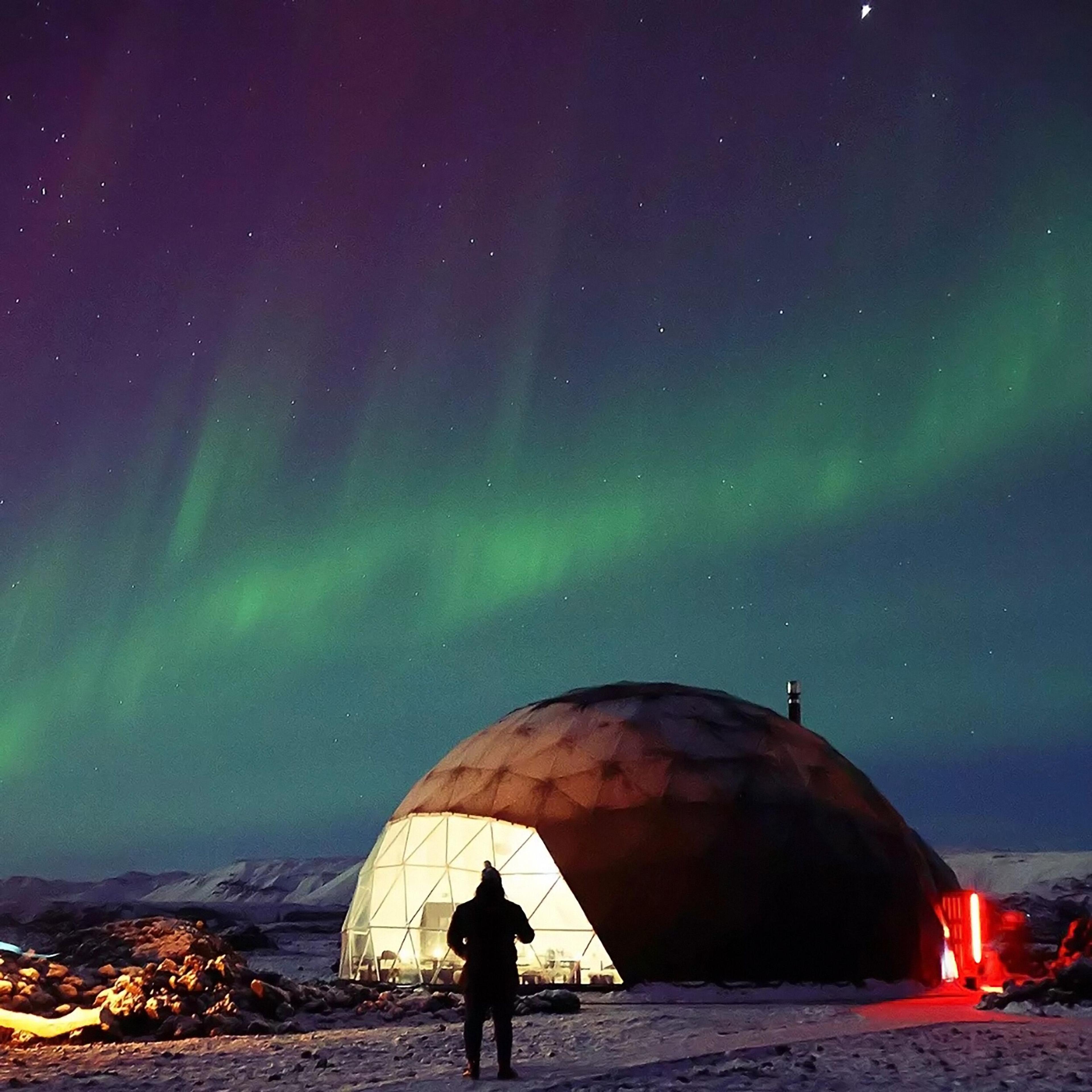 A geodesic dome at Aurora Base Camp in Iceland sits beneath vibrant northern lights, with a snowy winter scene enhancing the view.