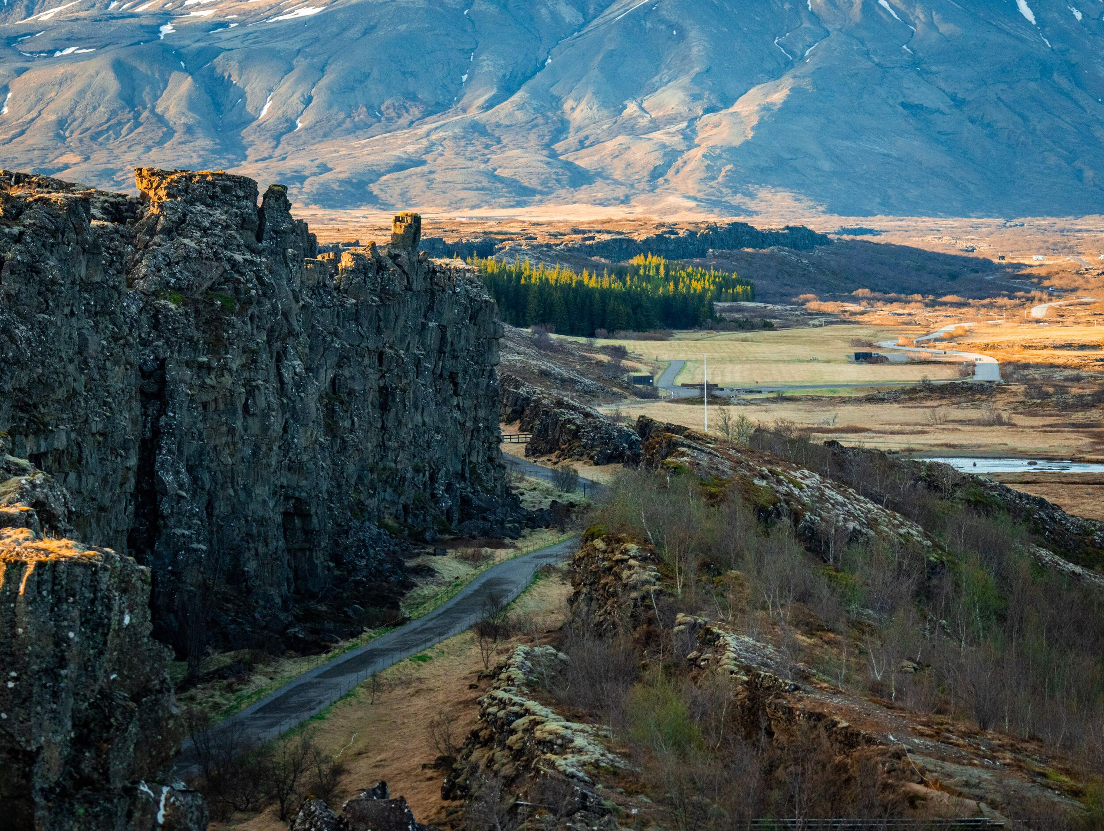 A winding path through a dramatic rocky gorge, opening to a wide valley with fields, a river, and distant mountains.