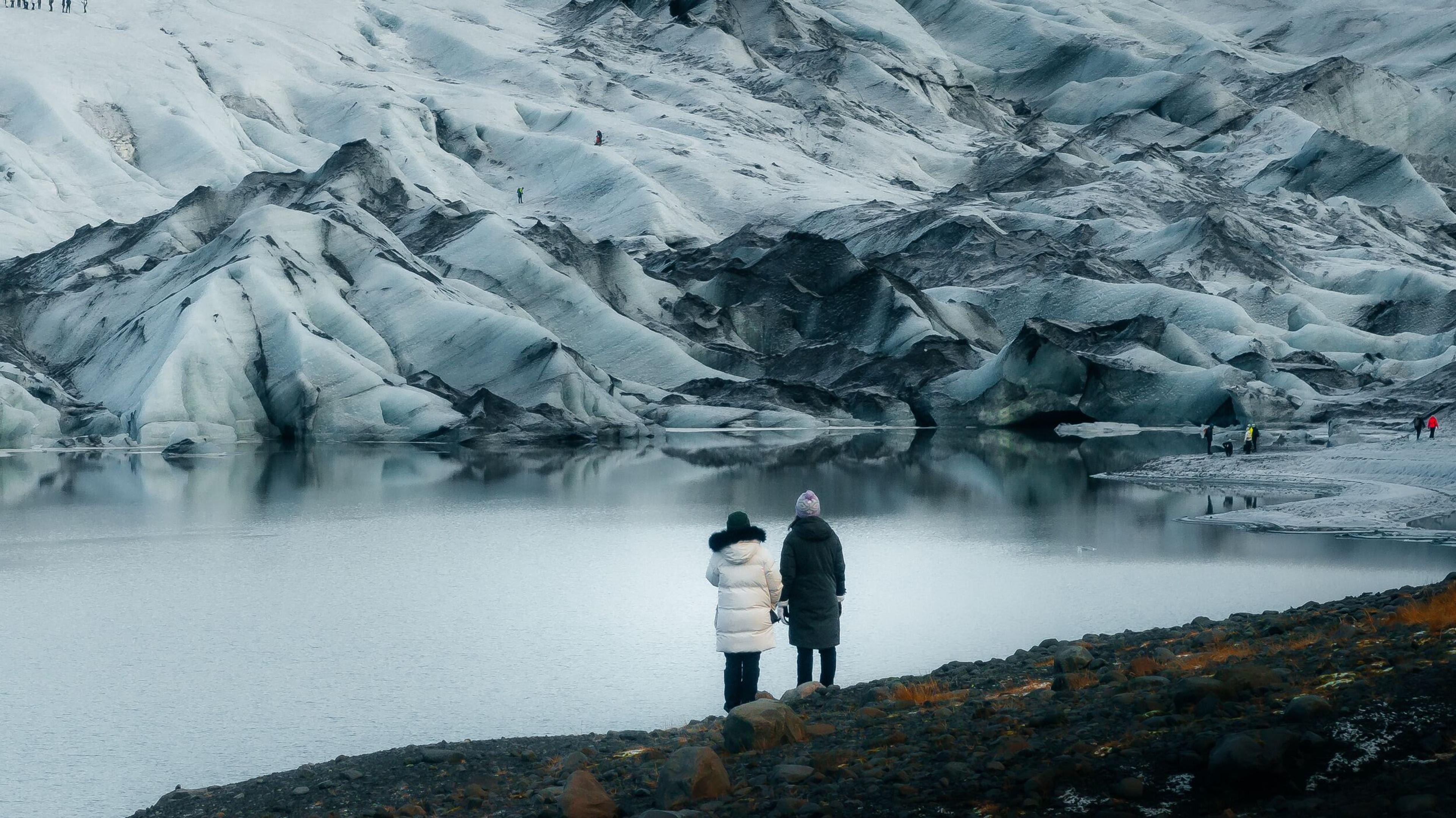 Two people stand by a glacial lagoon reflecting a vast glacier, with distant figures on the ice.