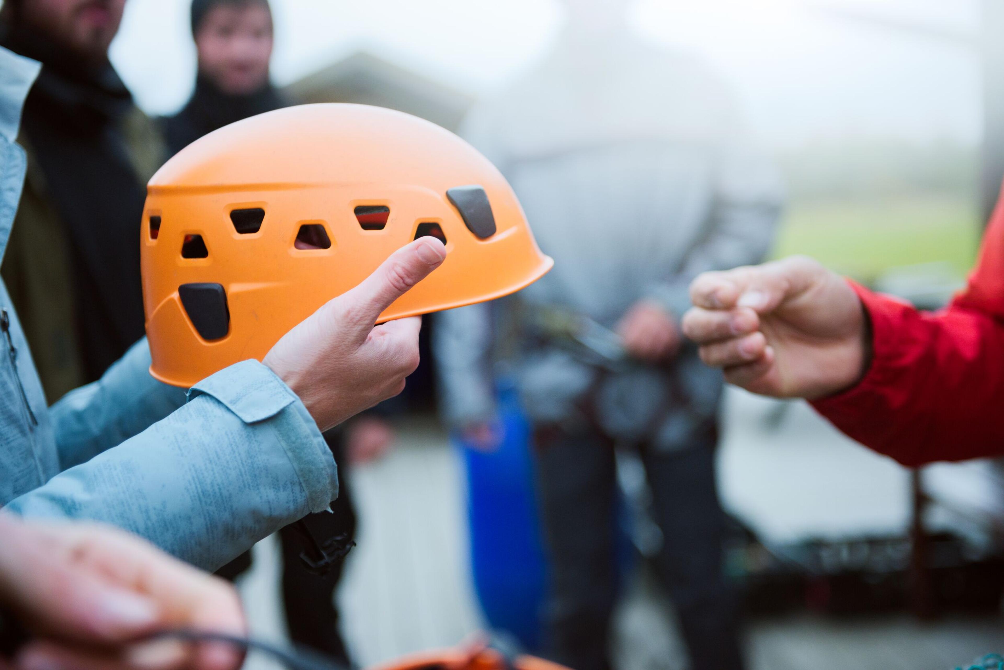 Person holding an orange safety helmet, preparing for an outdoor adventure with a group of people in the background.