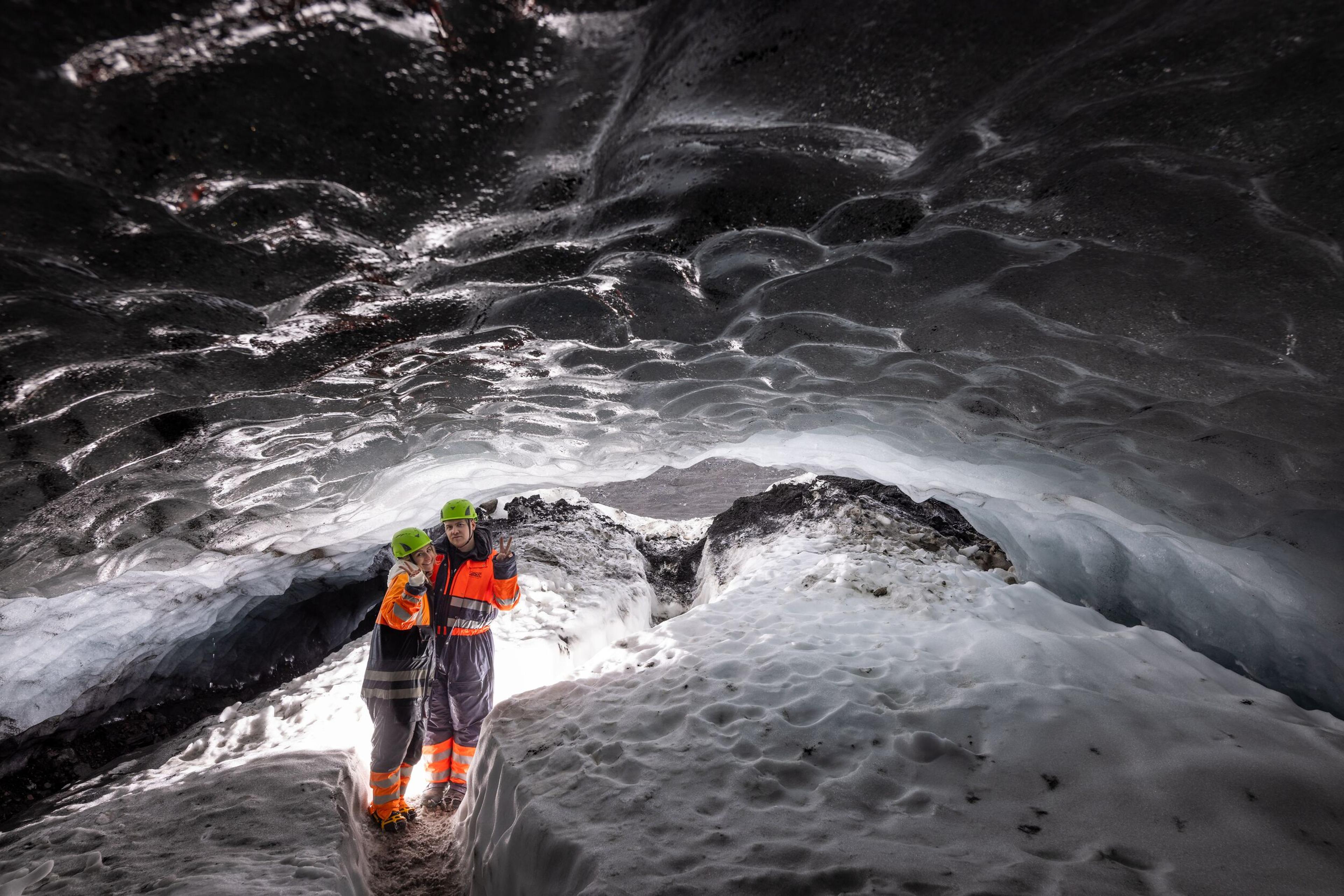 Two explorers in safety gear inside Askur ice cave, surrounded by glistening ice and snow with a textured ceiling.