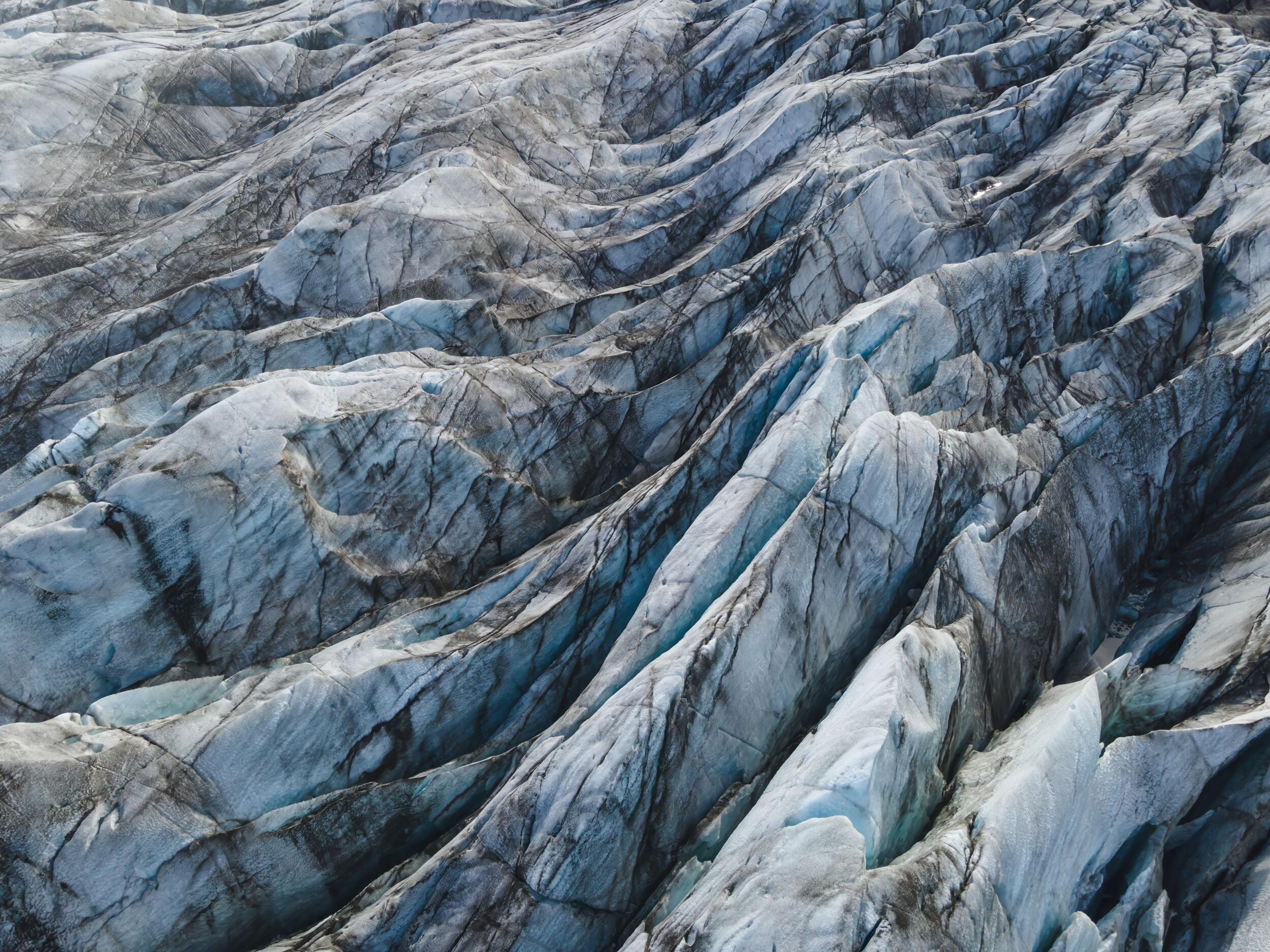 Close-up aerial view of a glacier with deep crevasses and a pattern of dark sediment lines, highlighting the dynamic and rugged texture of the ice.