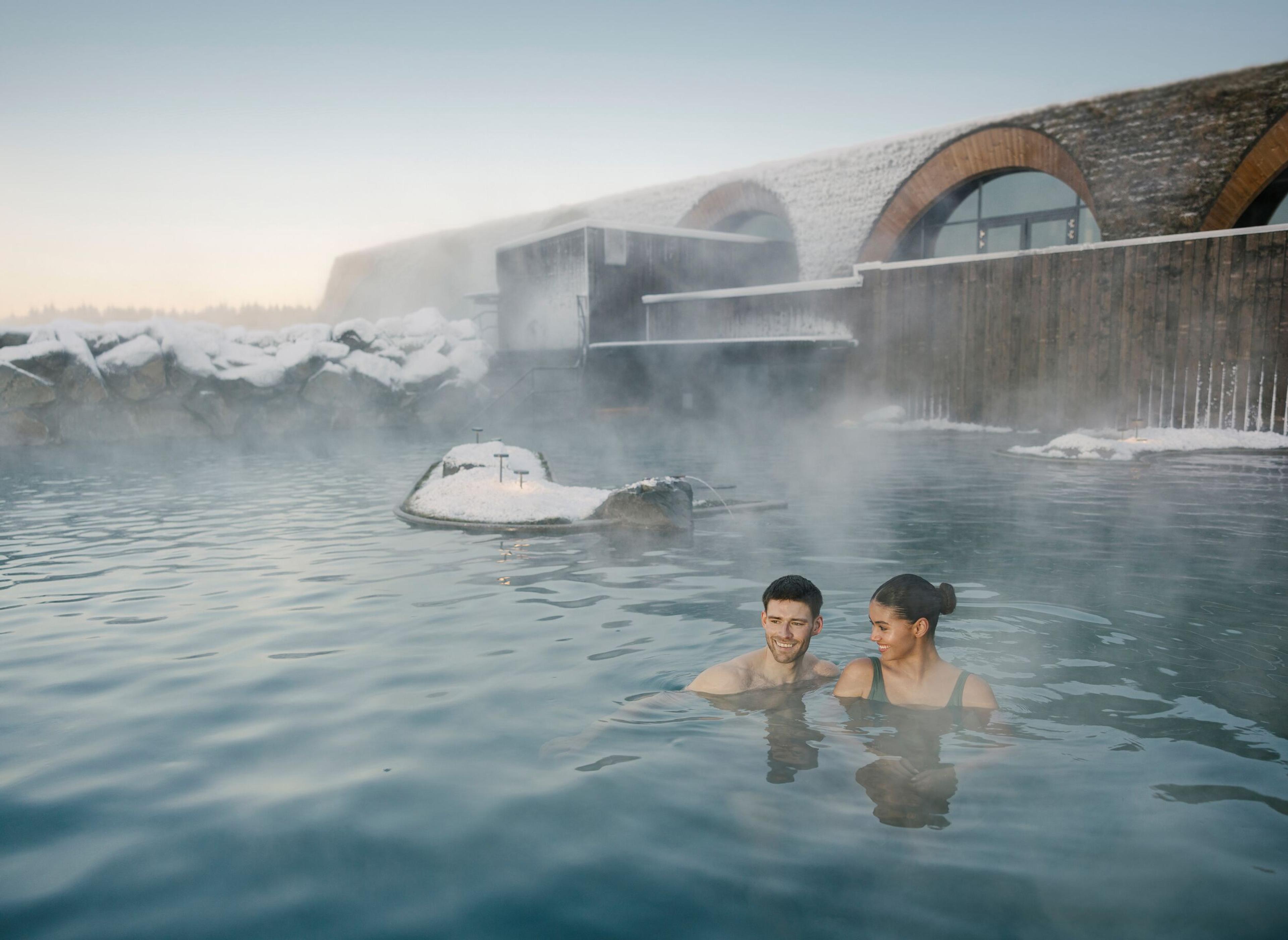 A couple relaxes in a steaming outdoor geothermal pool surrounded by snow and a modern building in winter.