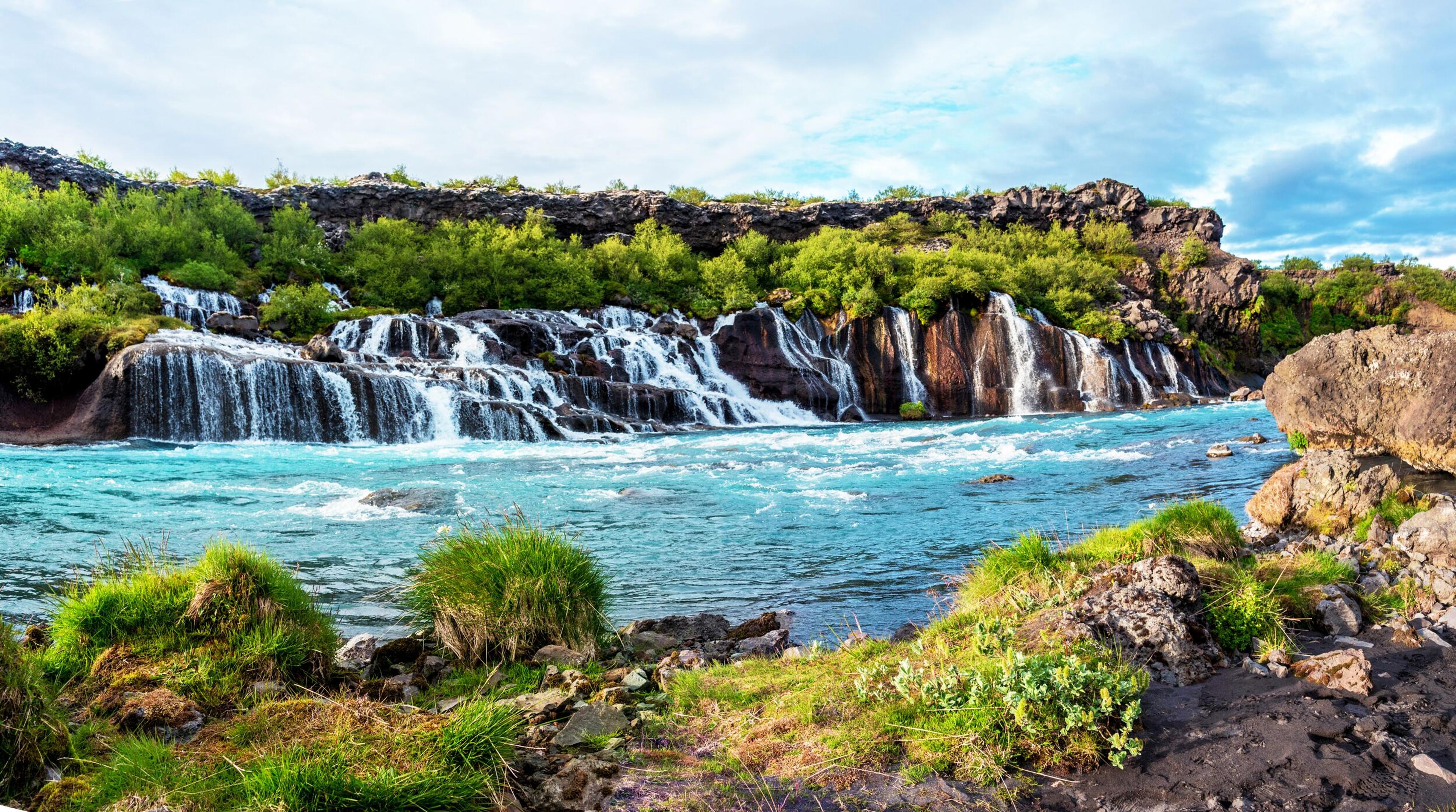 Turquoise river flowing past numerous small waterfalls cascading over dark rock and green foliage.