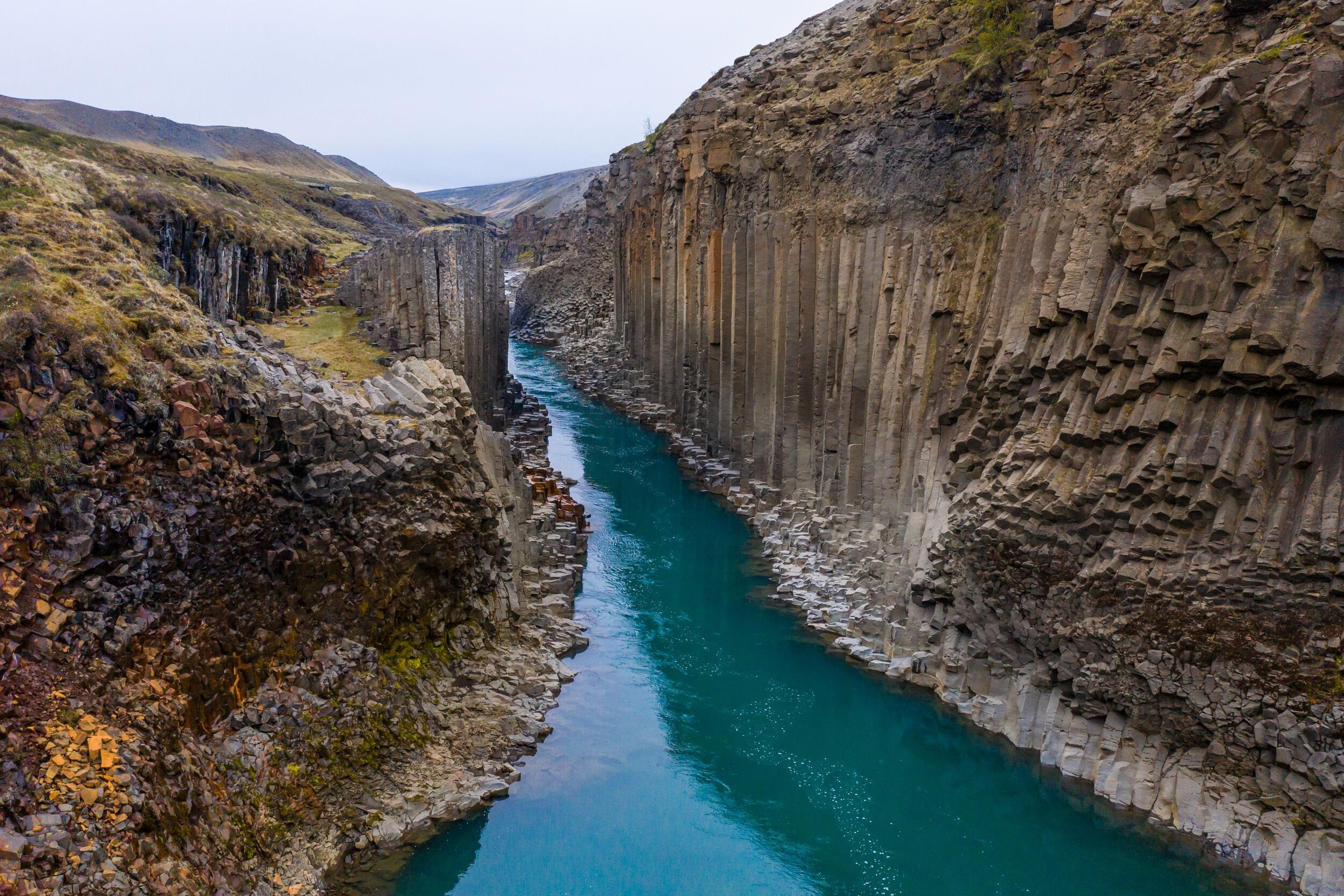 Aerial view of Stuðlagil Canyon in Iceland, showcasing a narrow river flowing between tall cliffs characterized by geometric basalt columns, with contrasting autumnal colors on the canyon's edges."