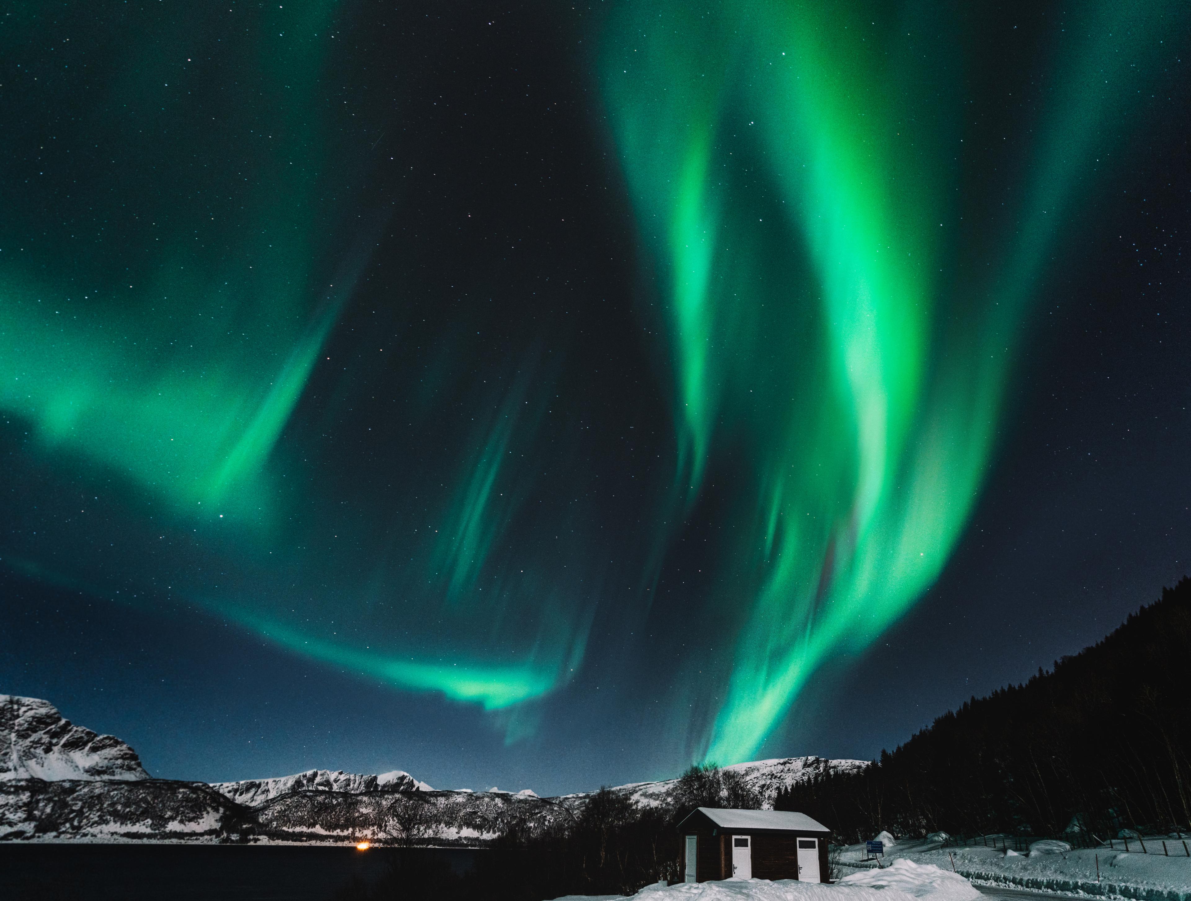 The Northern Lights sweep across the sky above a small cabin in a snowy landscape.