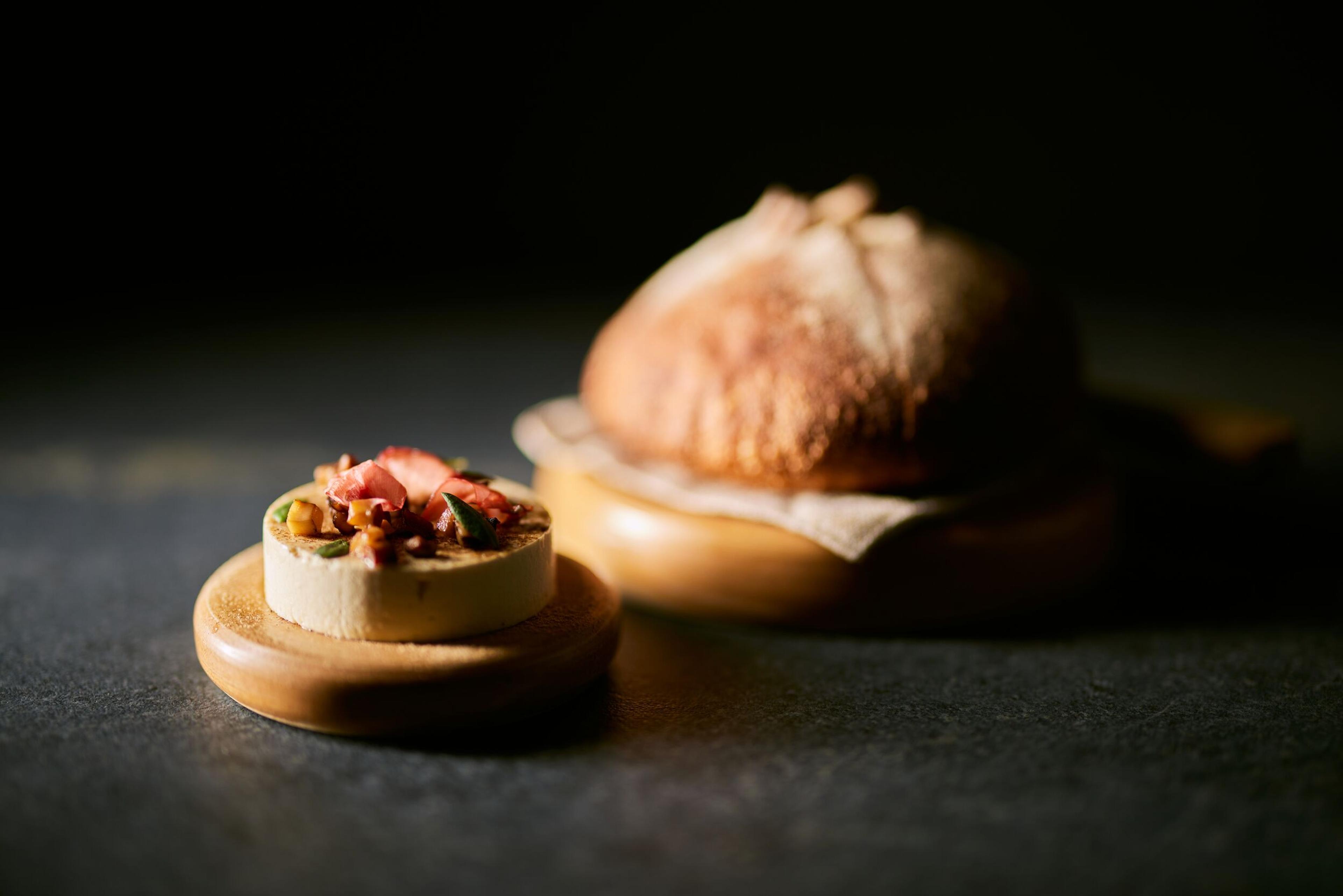 A small tart topped with strawberries and nuts, and a blurry rustic bread loaf, on wooden stands in dim light.
