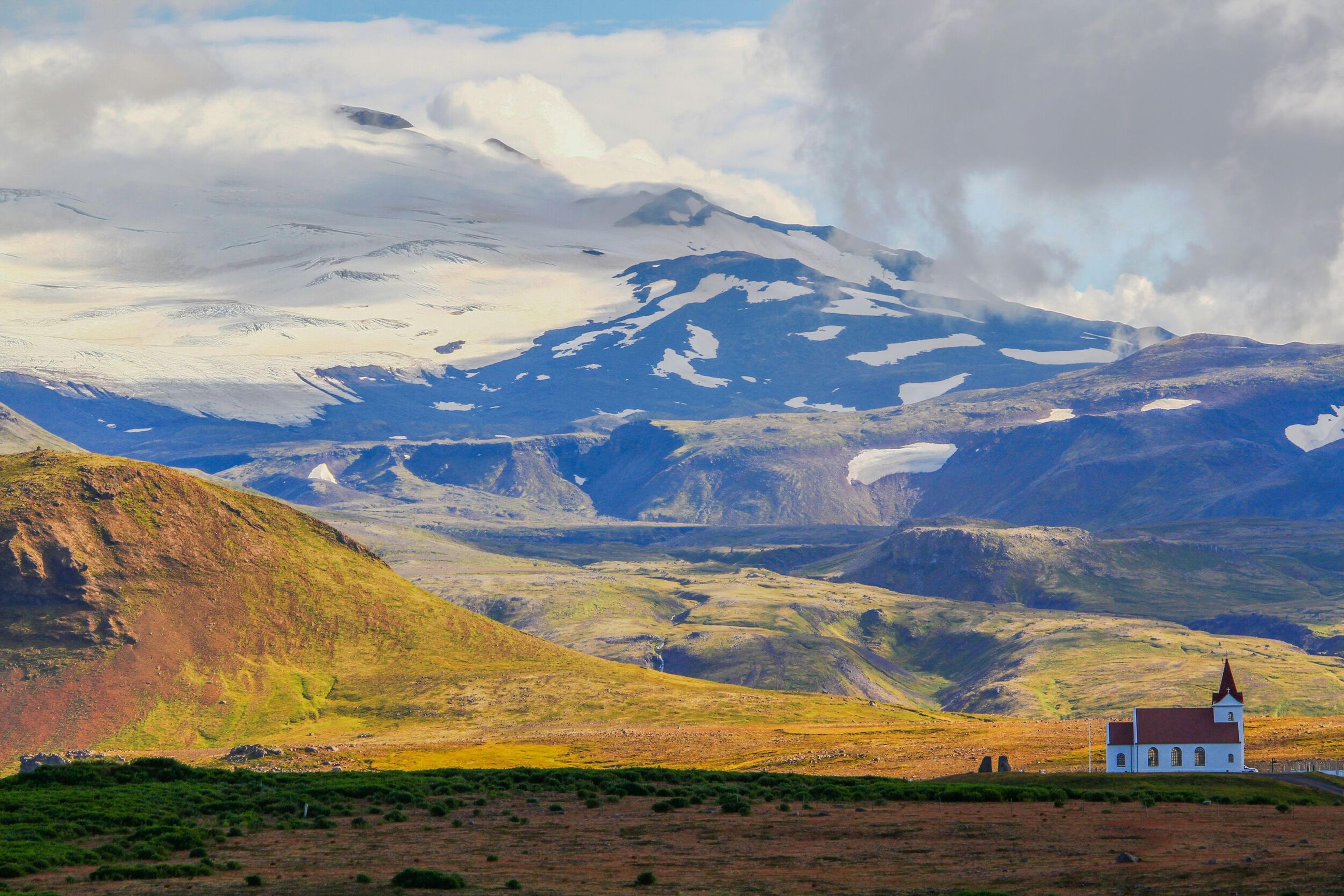 A lone church with a red roof sits in a vast landscape with lush green fields in the foreground and a large, snow-capped mountain range in the background under a cloudy sky.
