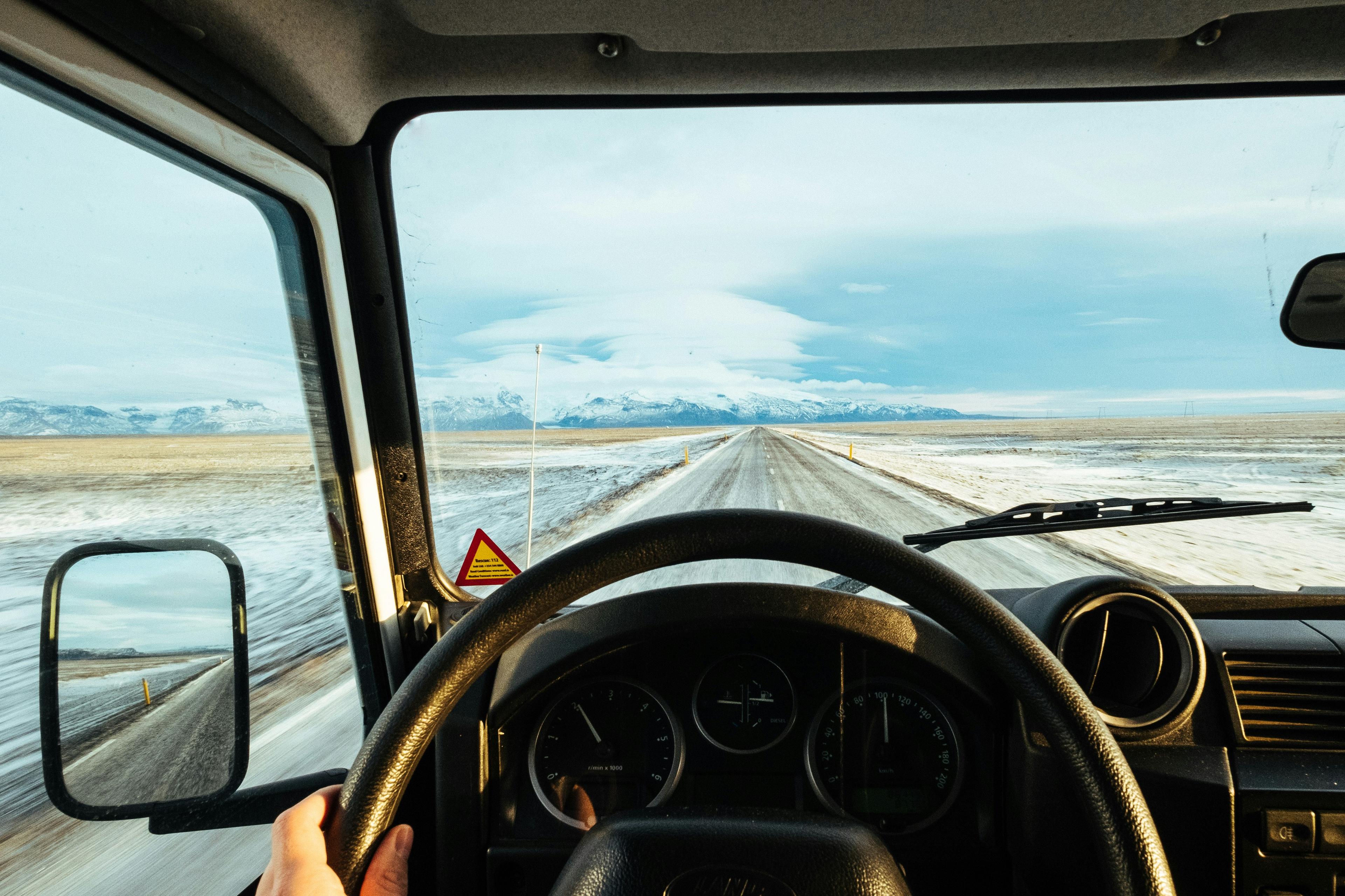 View from a vehicle's driver's perspective on a snowy road stretching into the distance with mountains and a clear sky ahead.