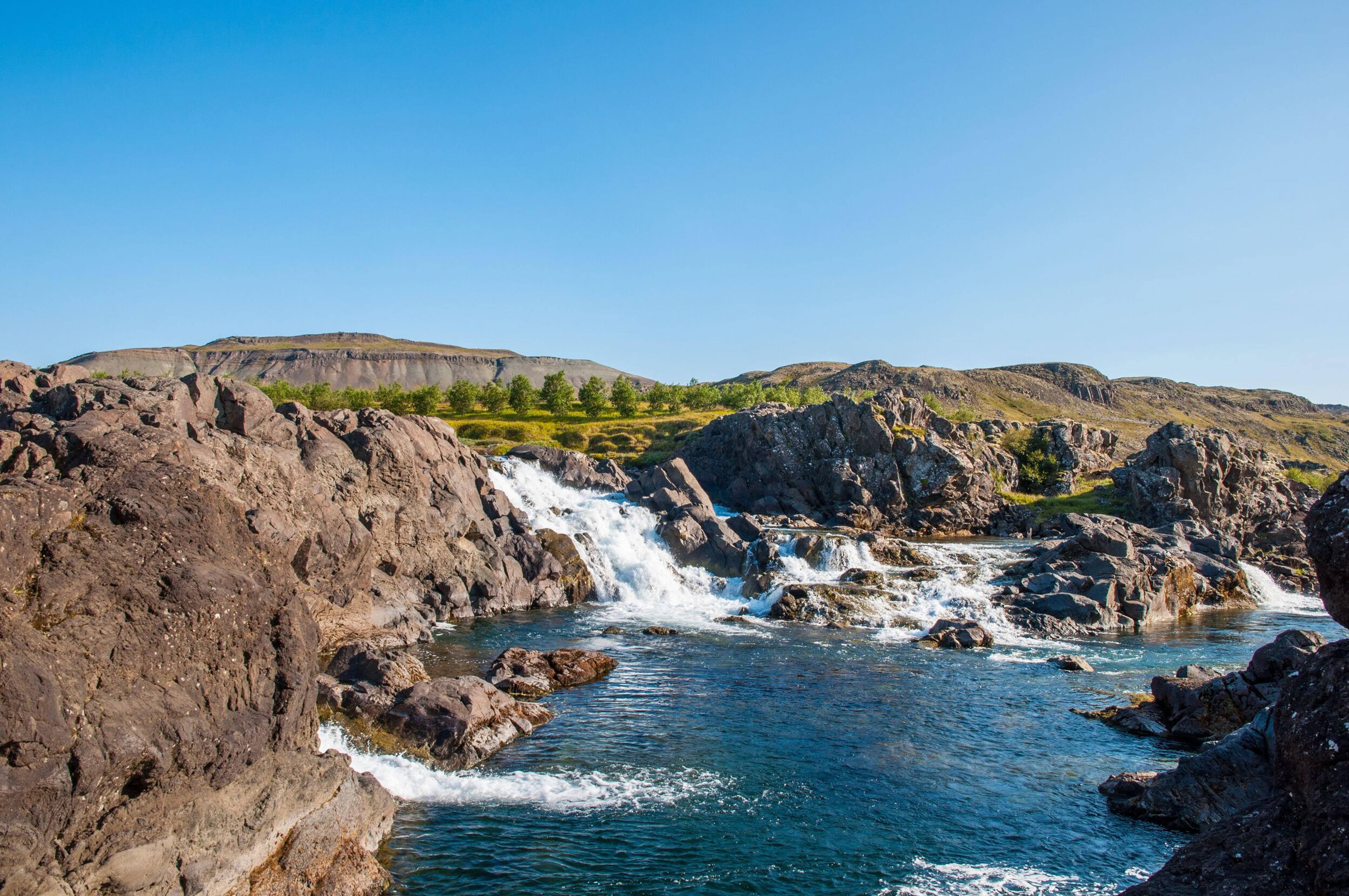 Scenic view of Glanni Waterfall in Iceland, surrounded by rugged volcanic rocks and lush greenery under a bright blue sky