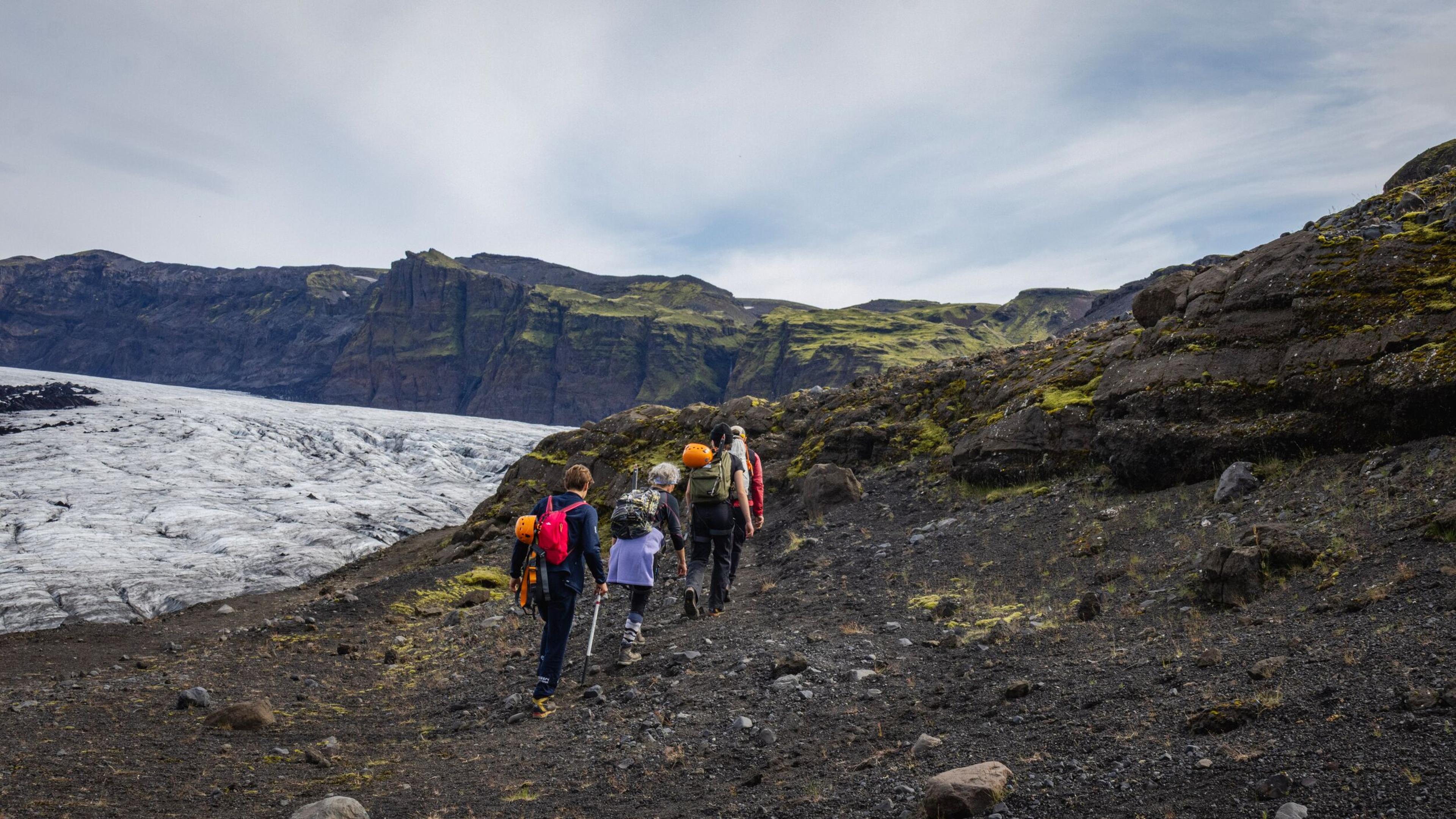 Hikers on a dark, rocky path overlooking a vast glacier and rugged mountains.