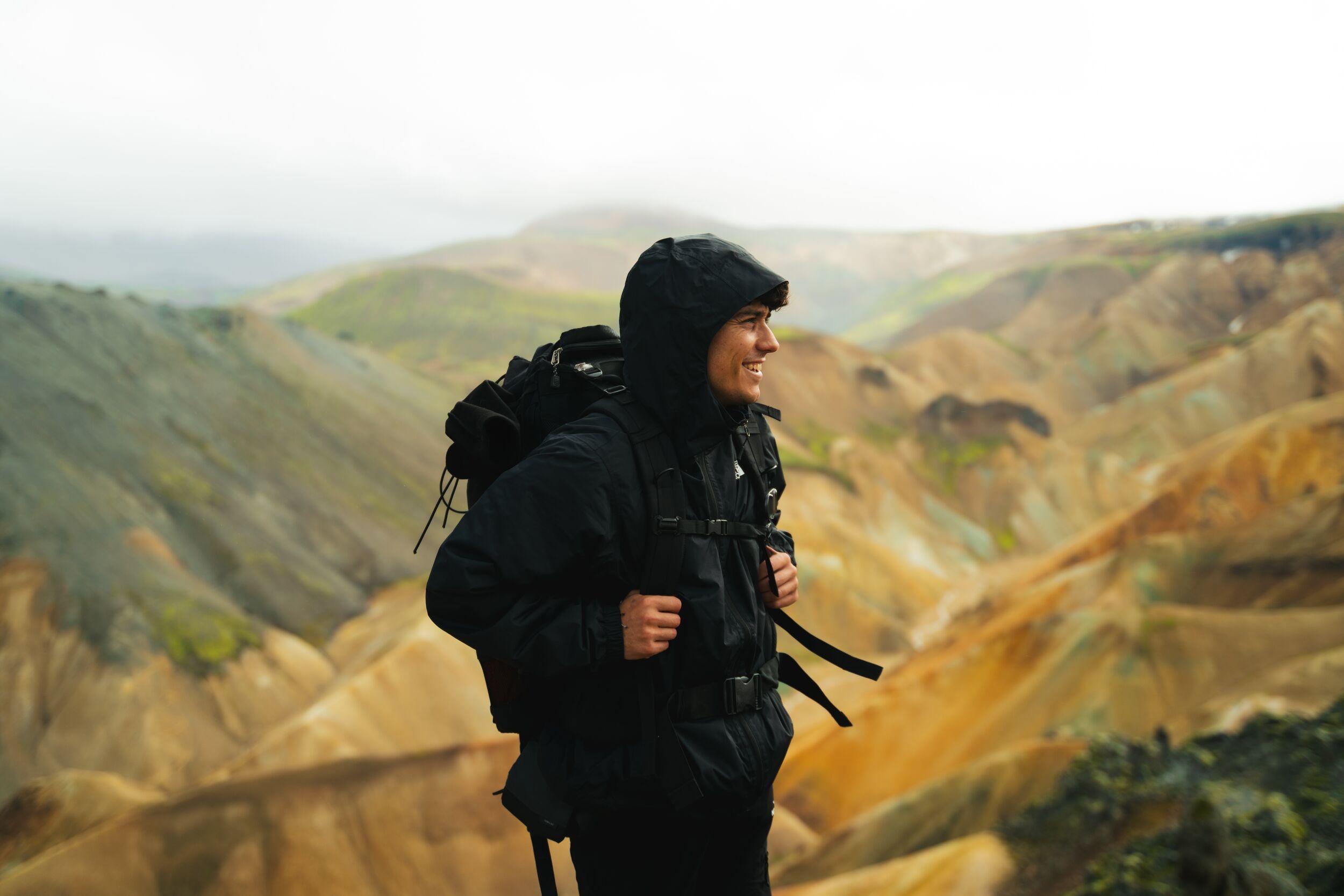 A smiling hiker in a black jacket and backpack looks over colorful, rolling mountains.