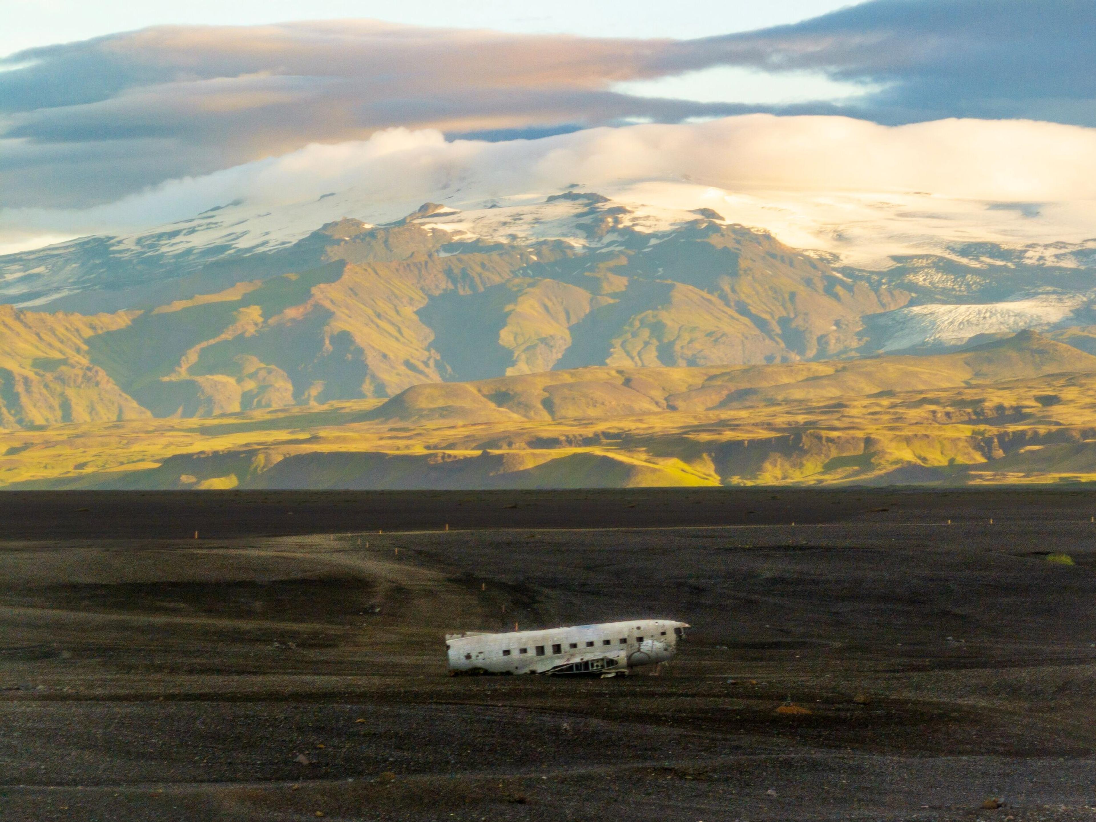 A white plane wreckage rests on a vast black sand plain, with distant snow-capped mountains under a warm, cloudy sky.