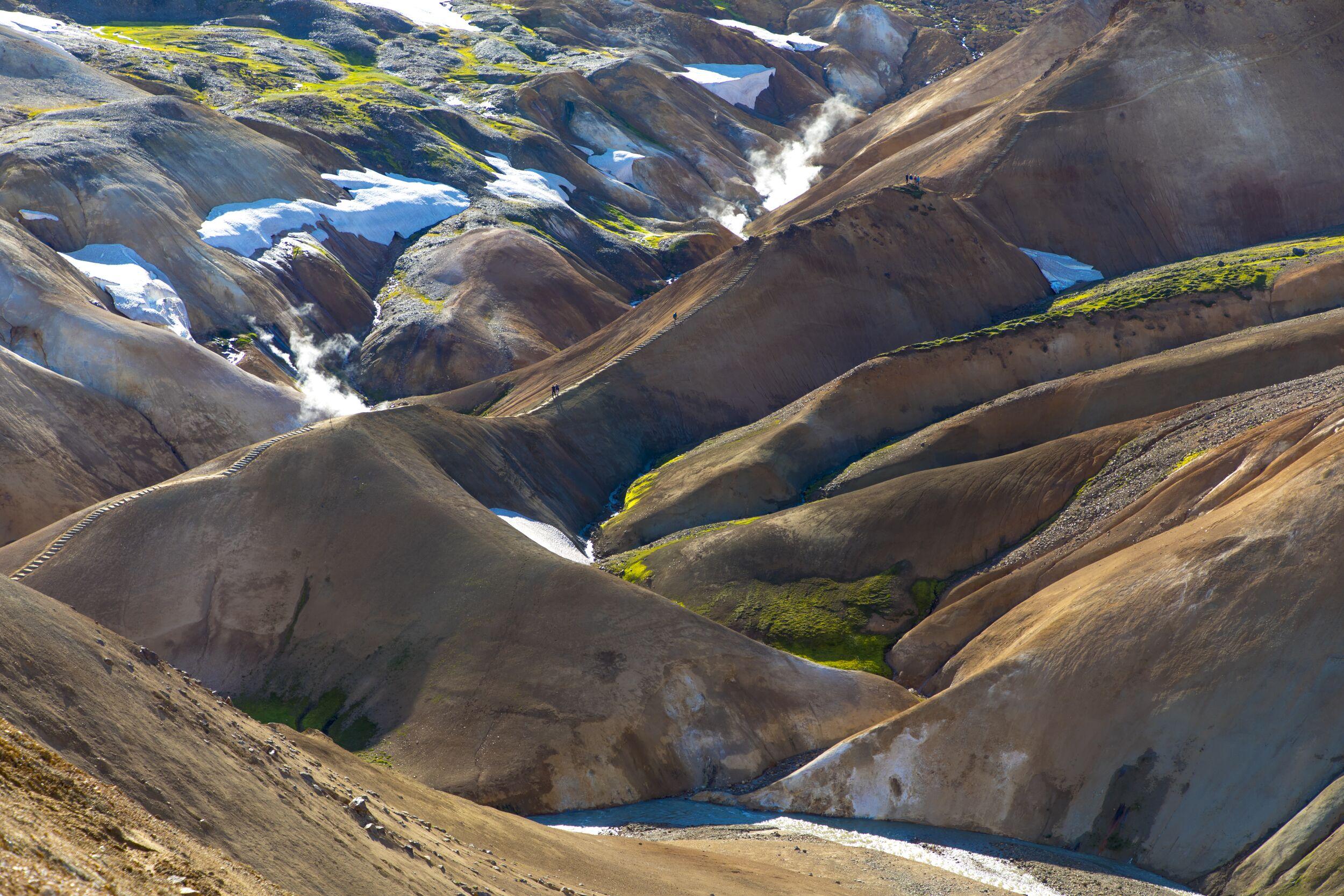 Colorful geothermal landscape with rolling hills, snow patches, and steam vents, with a riverbed below.