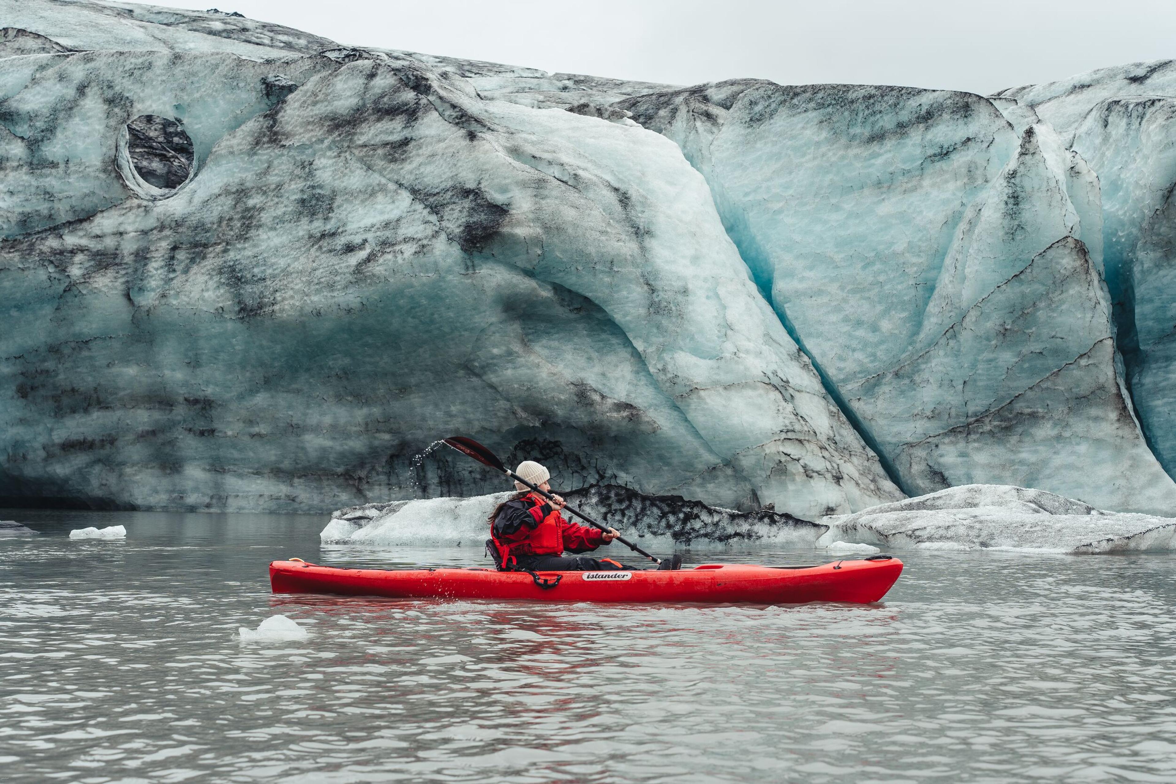 a man is paddling a red kayak in front of a large iceberg .