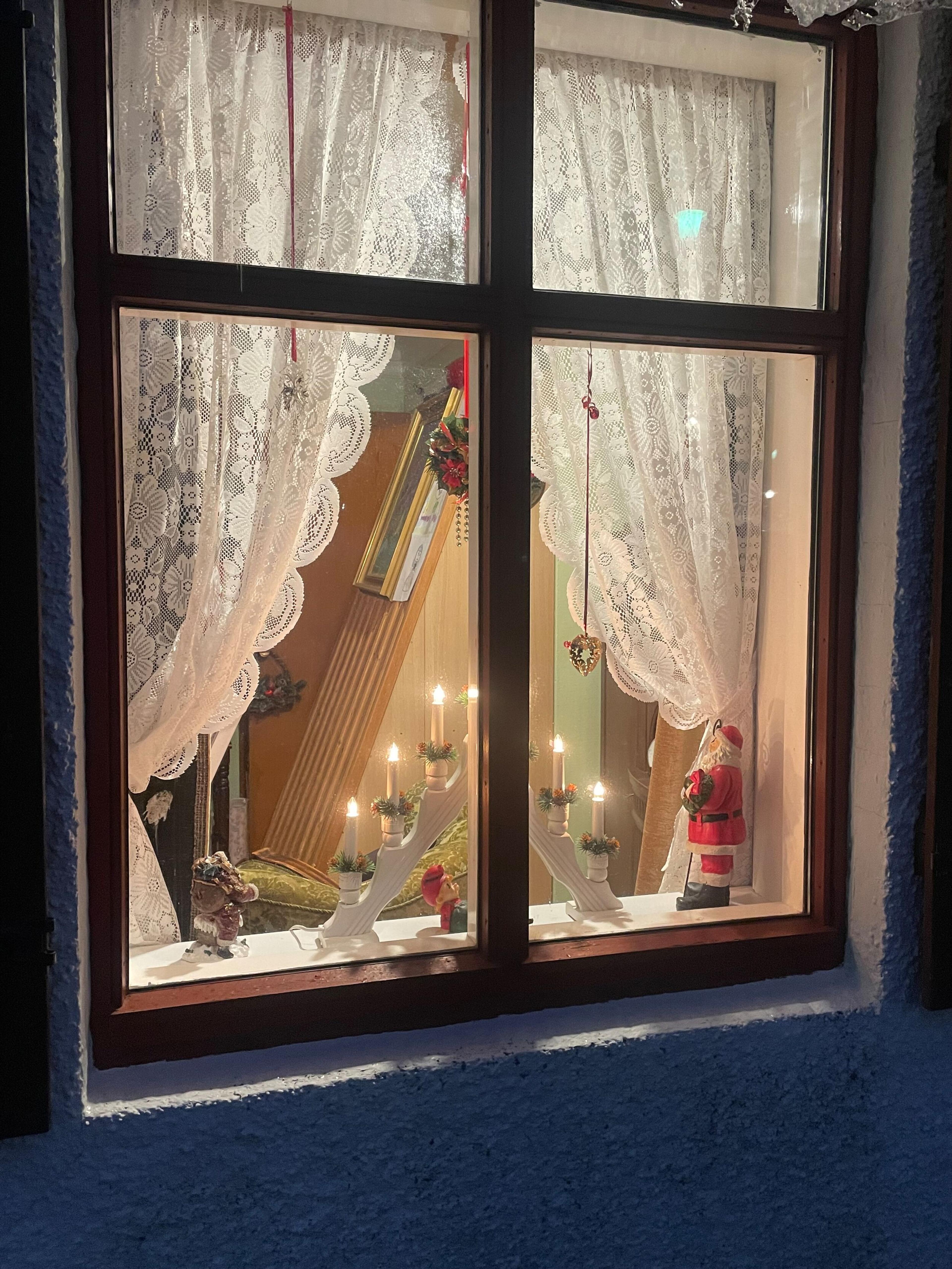A window with lace curtains displaying illuminated candle arches and a Santa figurine at night.