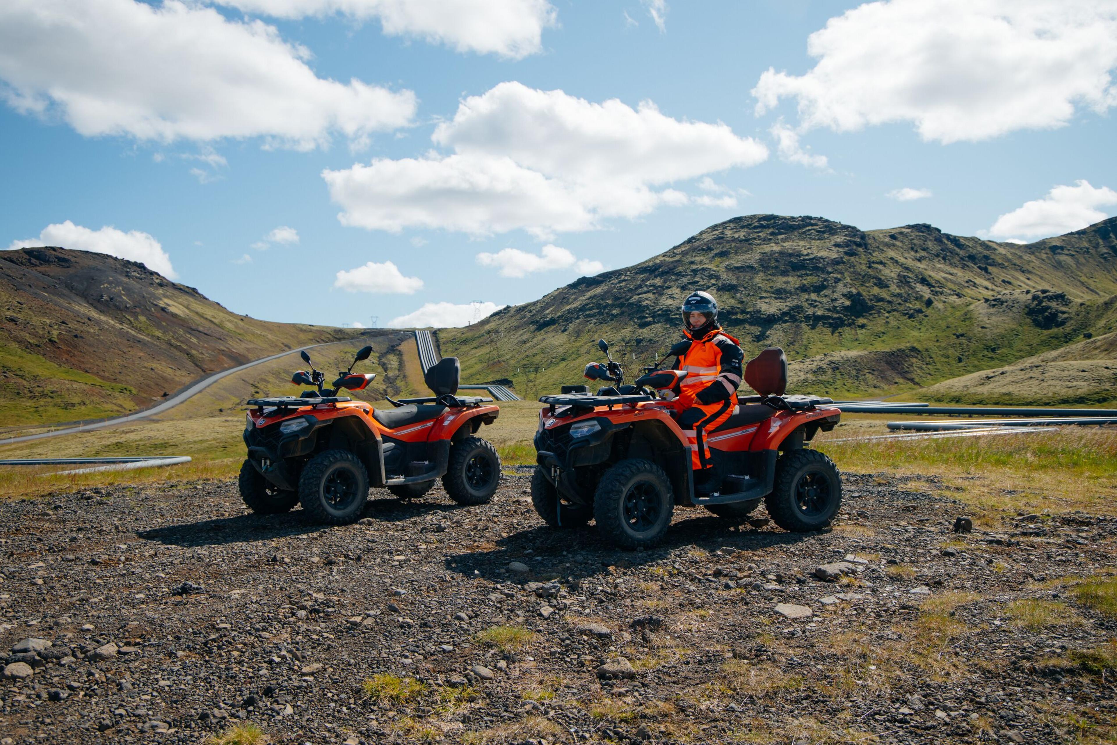 Two orange ATVs on a gravel trail, one with a rider in an orange jacket and helmet, backed by mountains and a winding road.