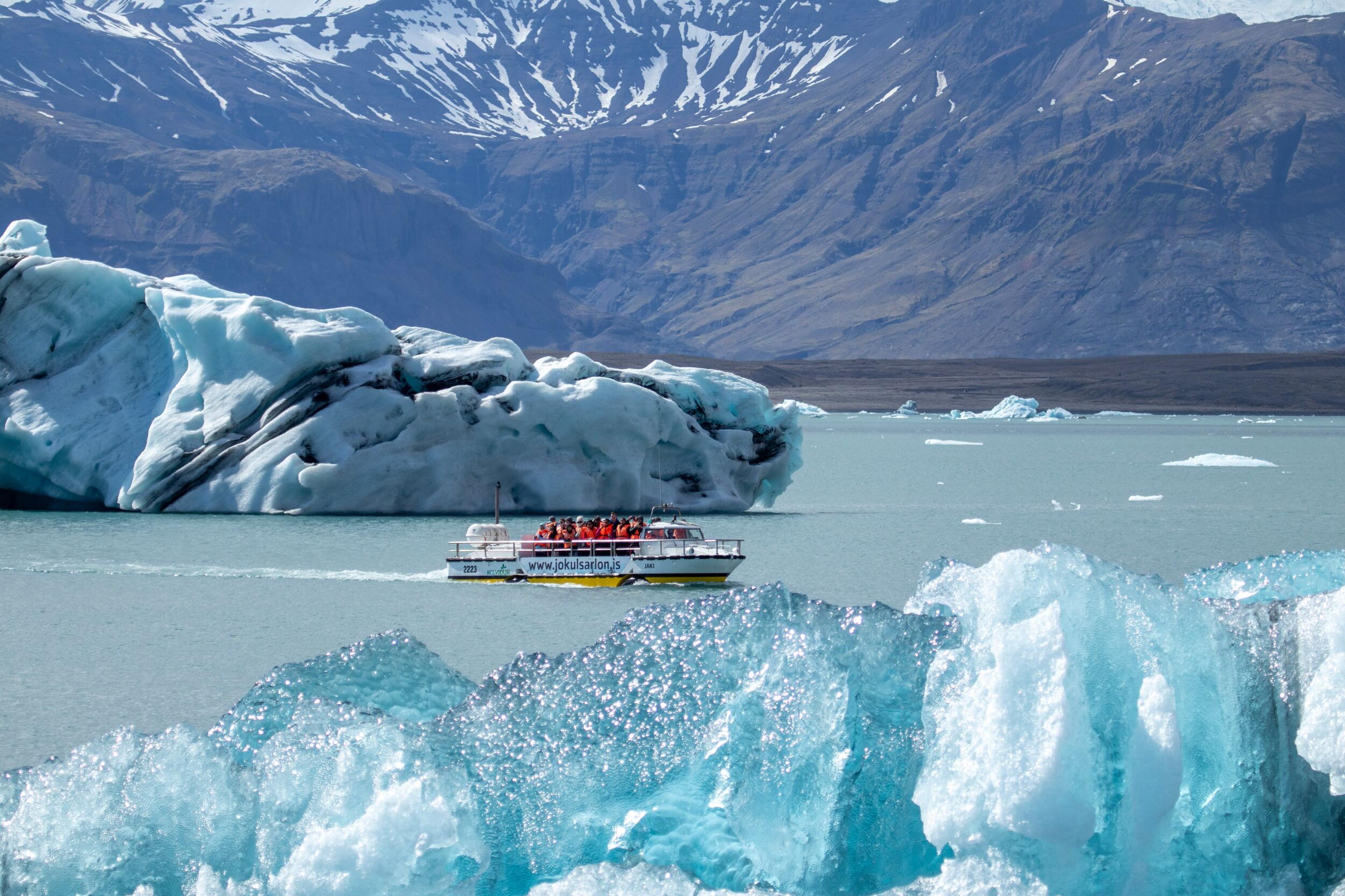 A tour boat navigates a glacial lagoon amidst icebergs, with snow-capped mountains in the distance.