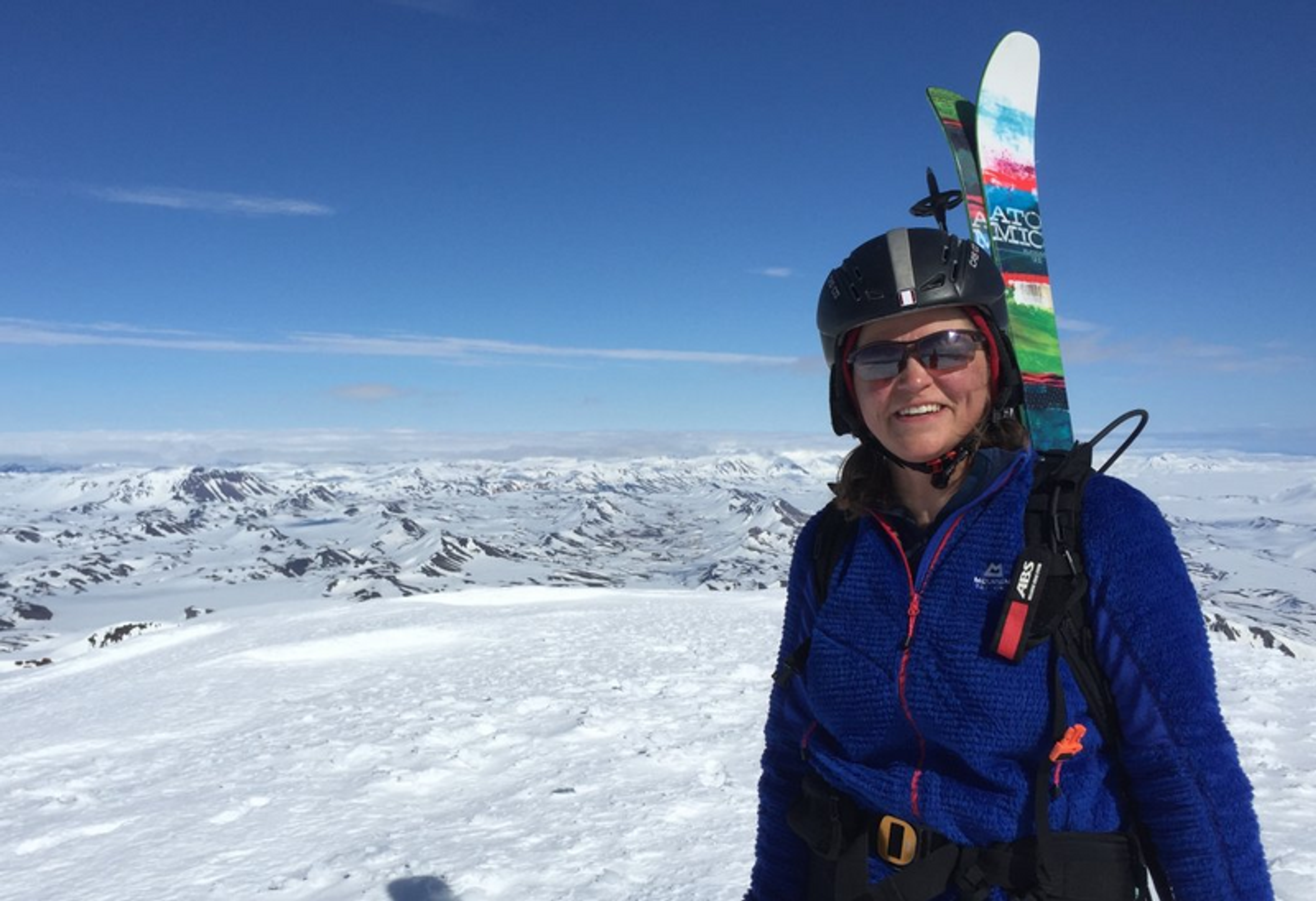 A smiling individual in a blue jacket and ski helmet, with skis attached to their backpack, stands atop a snowy landscape with a panoramic view of mountainous terrain under a clear blue sky.