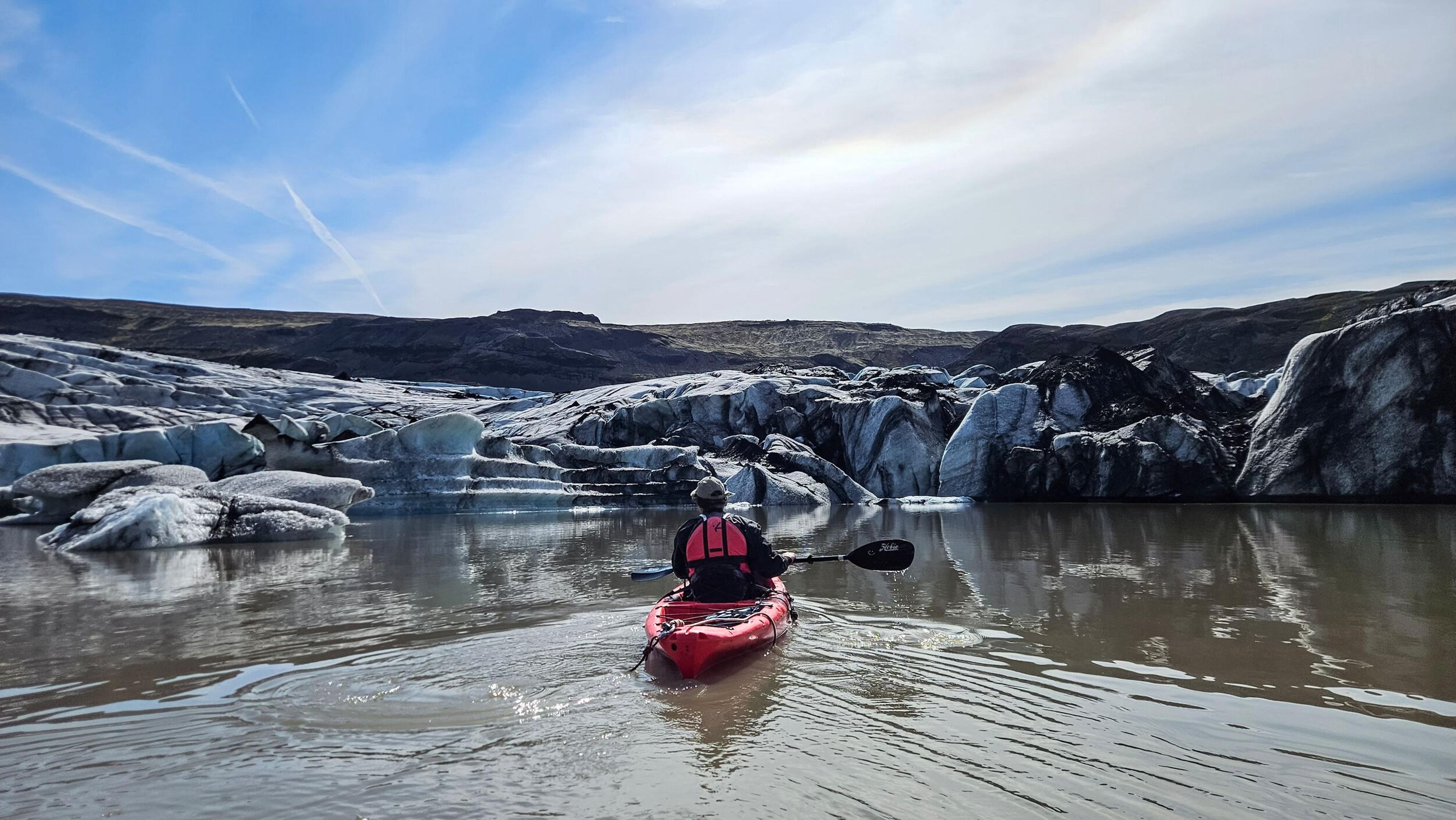 A person in a red kayak paddles on a glacial lagoon surrounded by ice formations and mountains under a blue sky.