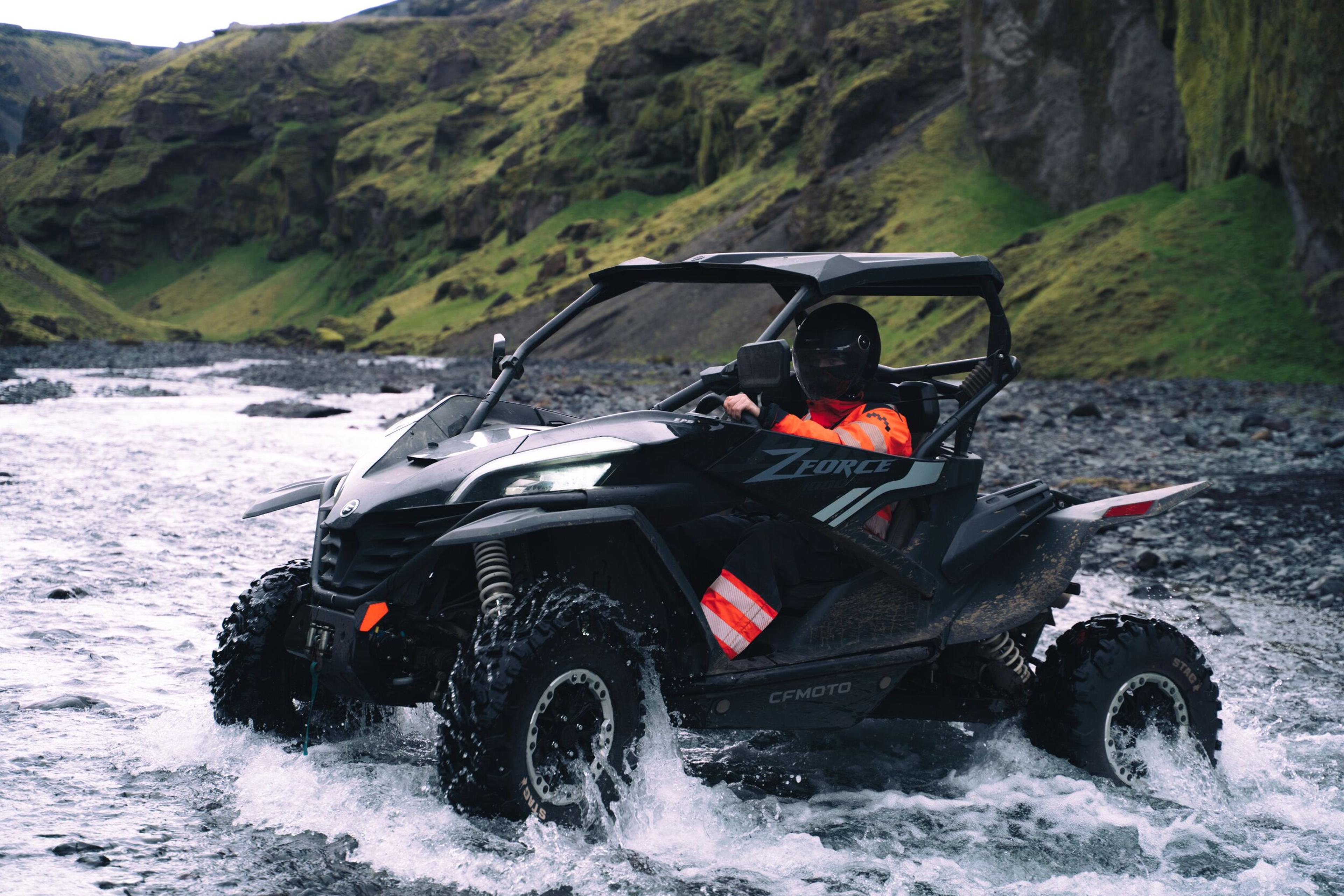 Black UTV driven by a person in a helmet and orange jacket splashes through a river with green hills in the background.