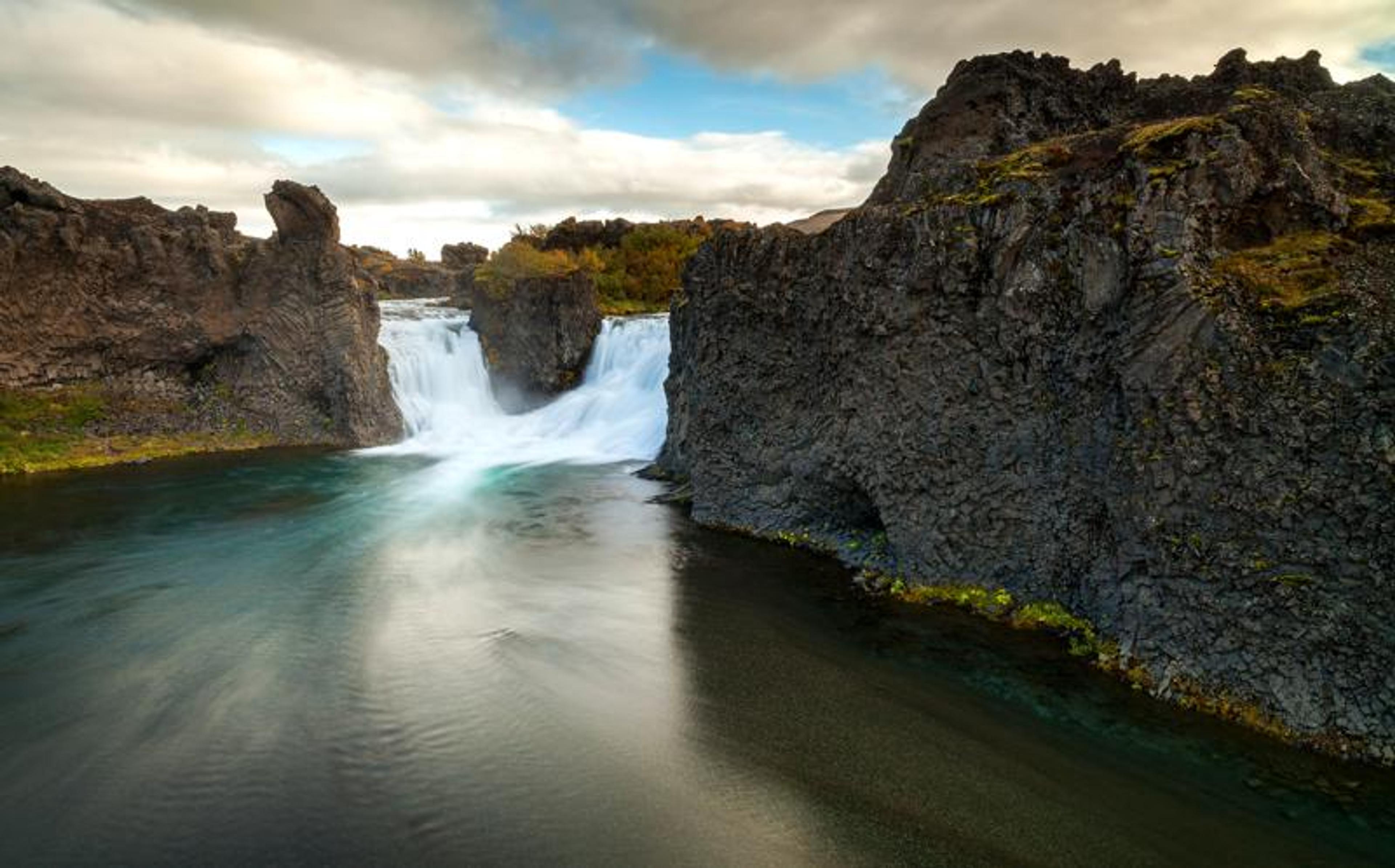 Wide waterfall between dark volcanic rock formations emptying into a river.