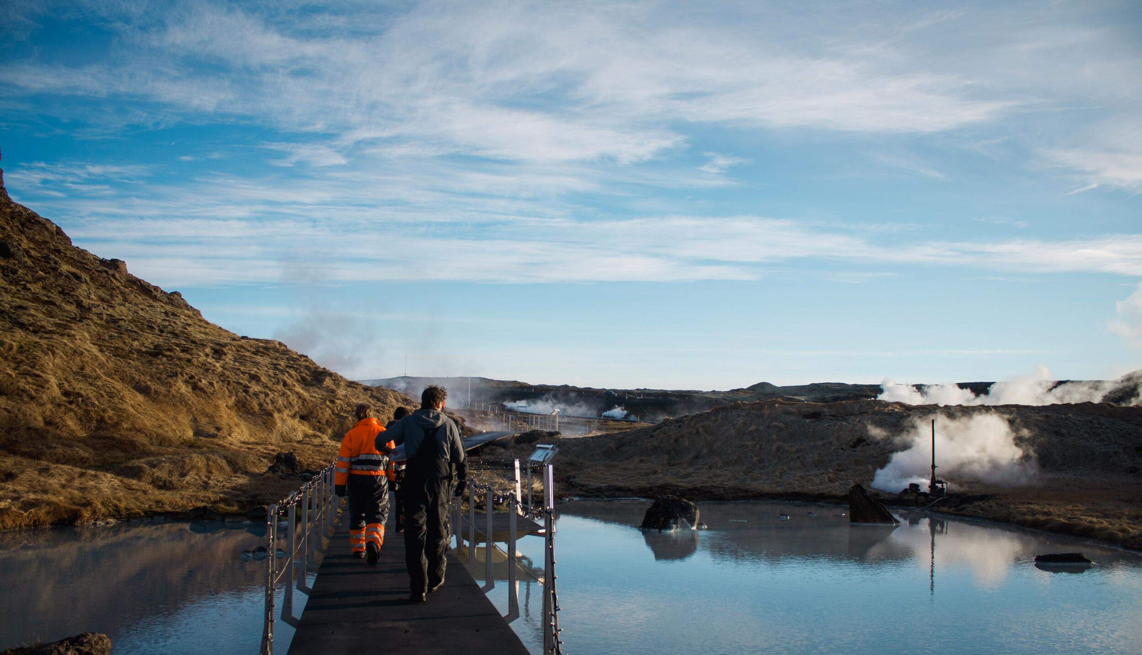 Three people walk on a boardwalk over a steaming geothermal pool surrounded by rocky terrain under a blue sky.