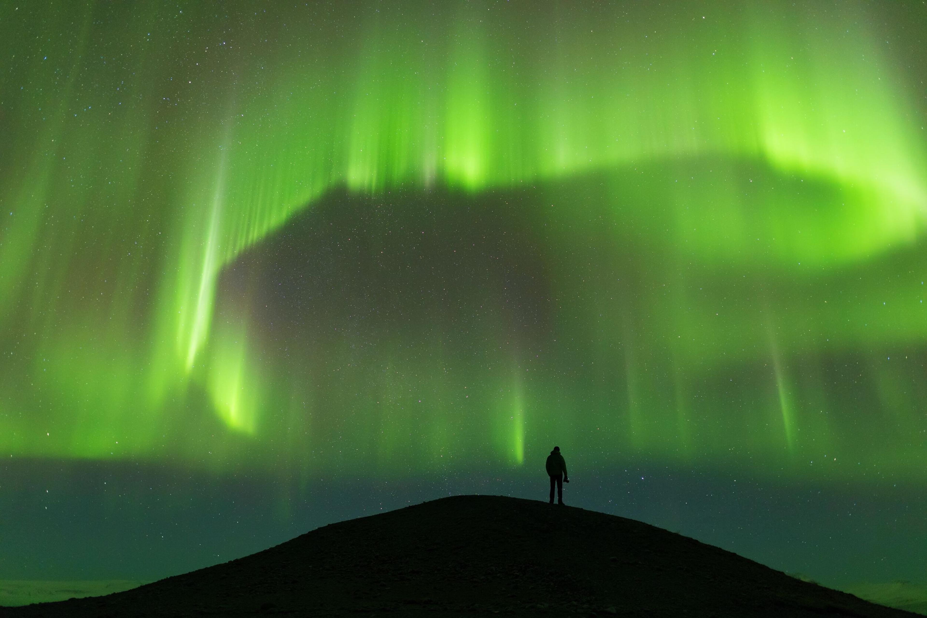 A lone figure stands silhouetted on a dark hill under vibrant green aurora borealis.
