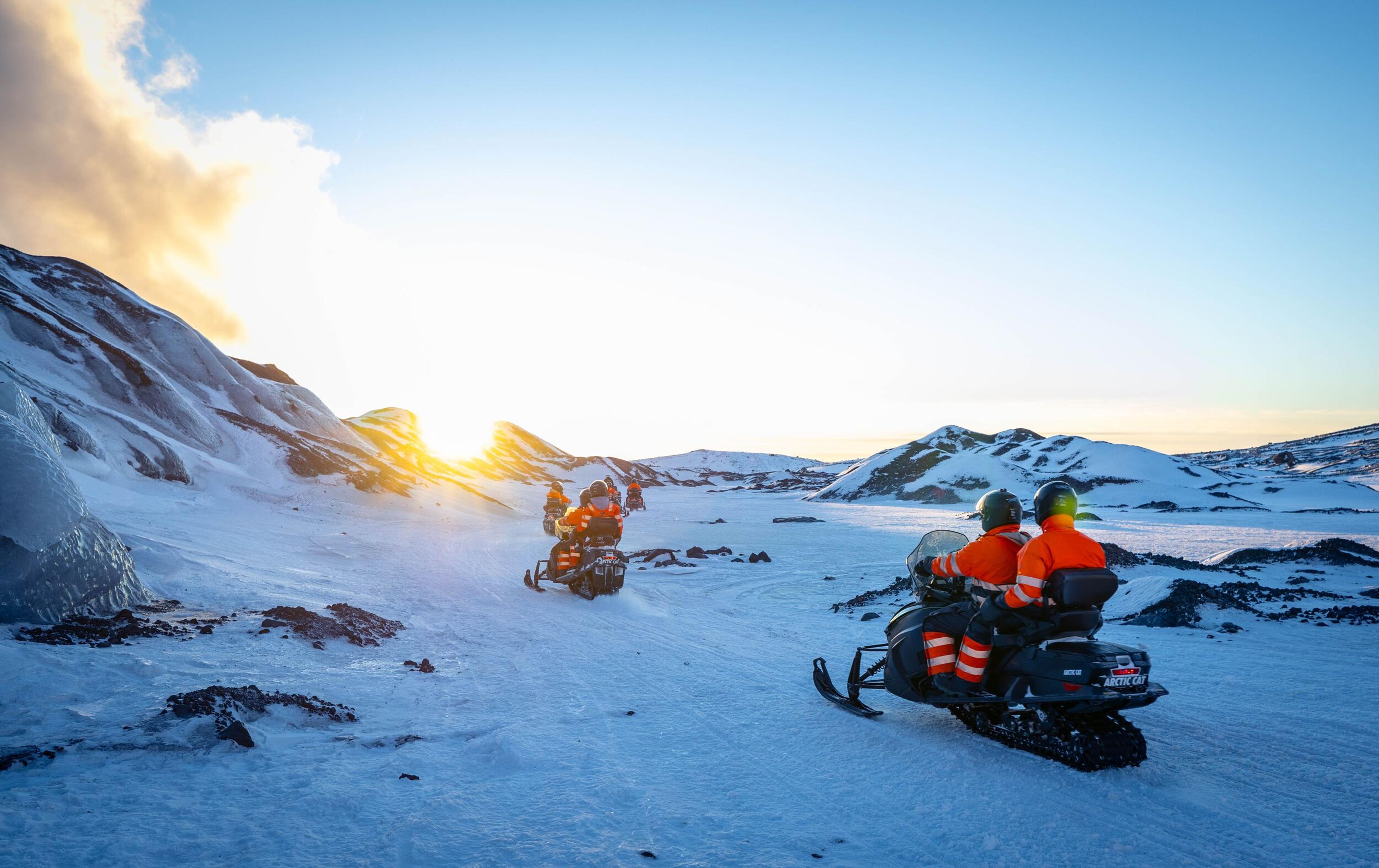 A group of adventurers on snowmobiles enjoy a bright day amidst a snowy landscape with rolling hills and a clear blue sky.