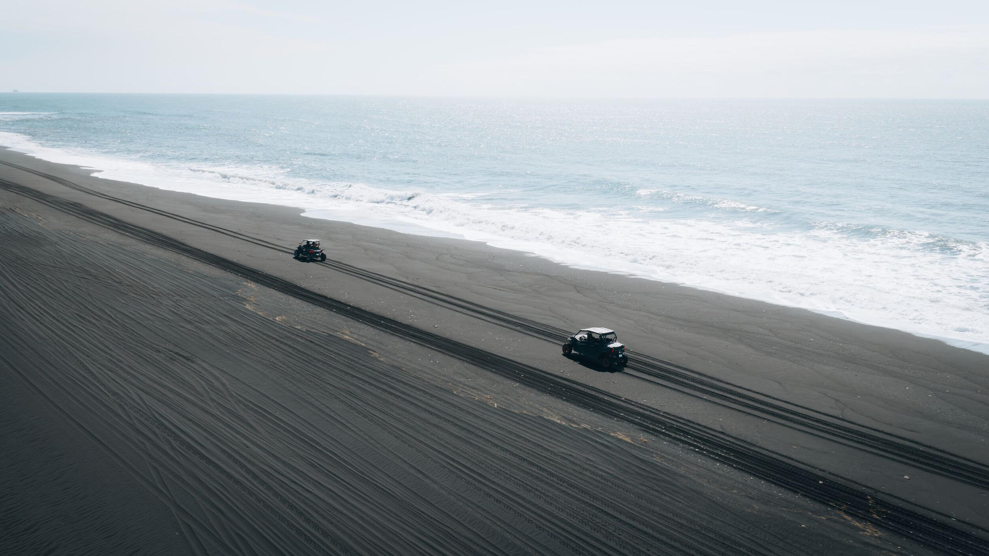 Two dark vehicles driving on a black sand beach next to the ocean.
