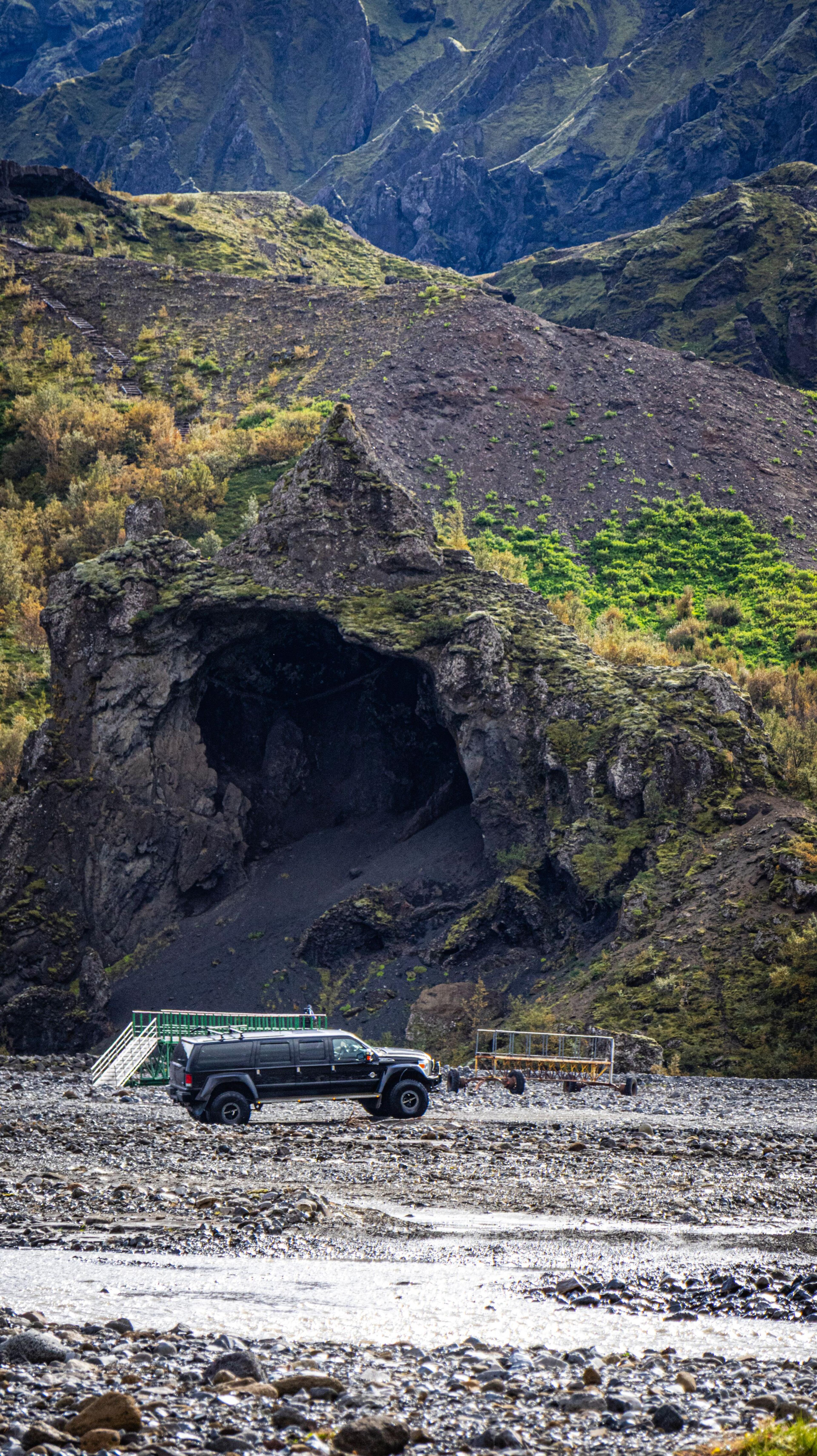A black off-road vehicle parked on a rocky riverbed in front of a large cave entrance in a mossy cliff, with steep mountains in the background.