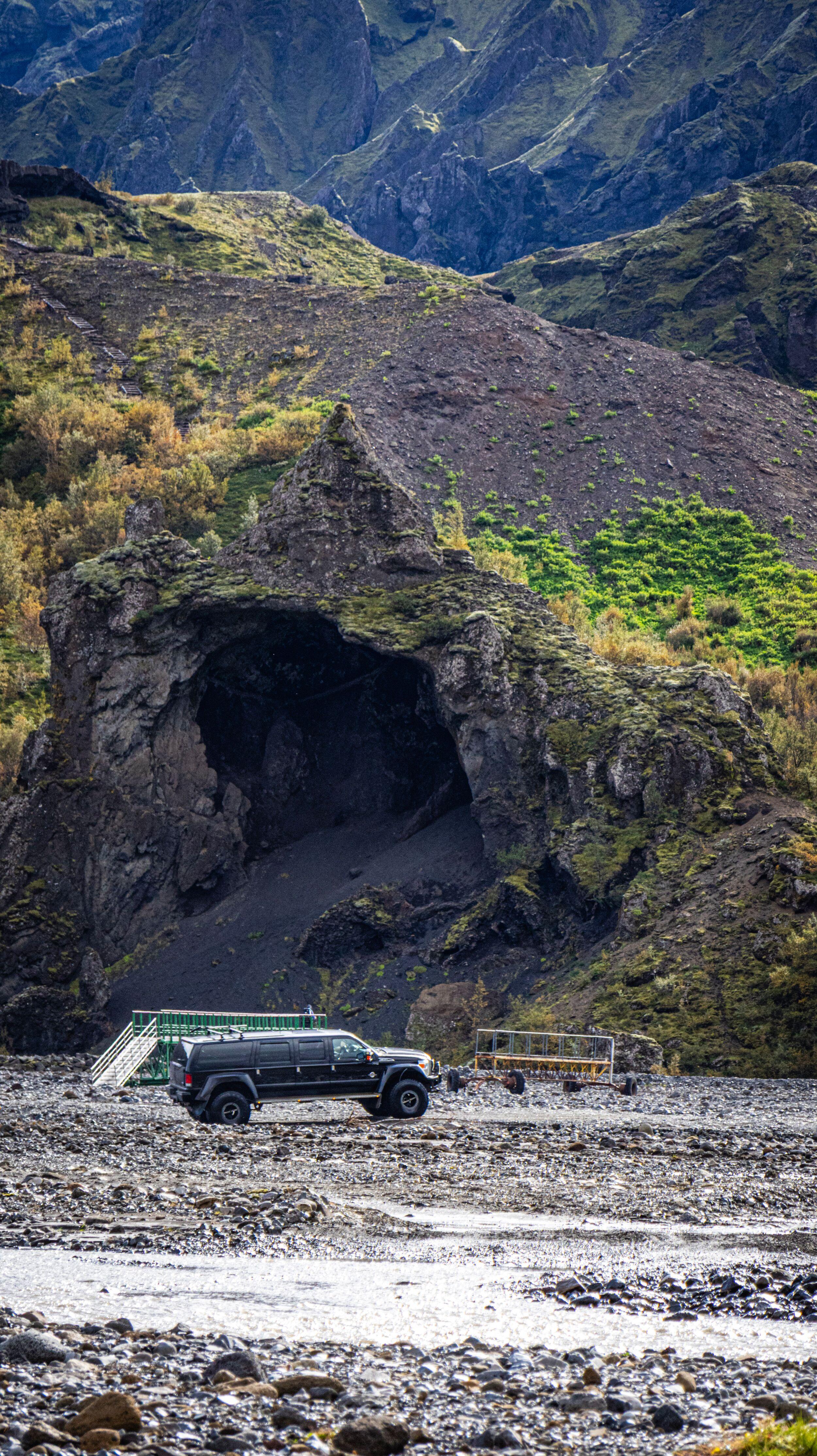 A black off-road vehicle parked on a rocky riverbed in front of a large cave entrance in a mossy cliff, with steep mountains in the background.