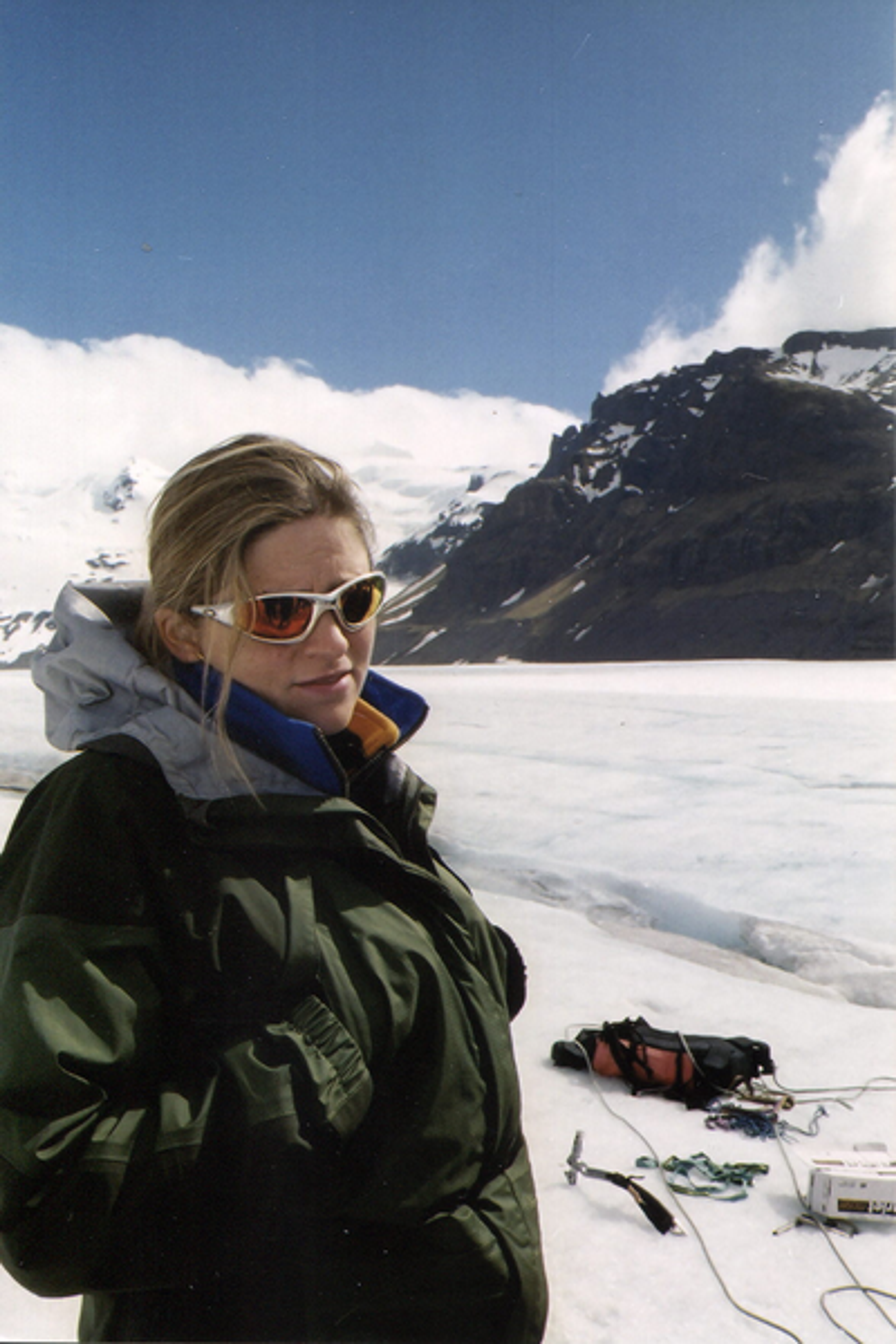 A person wearing sunglasses and a winter jacket stands in the foreground with a vast expanse of ice and snow stretching out behind them, under a mountainous horizon and blue sky. They appear equipped for a cold environment, with a radio and equipment visible on the icy ground.