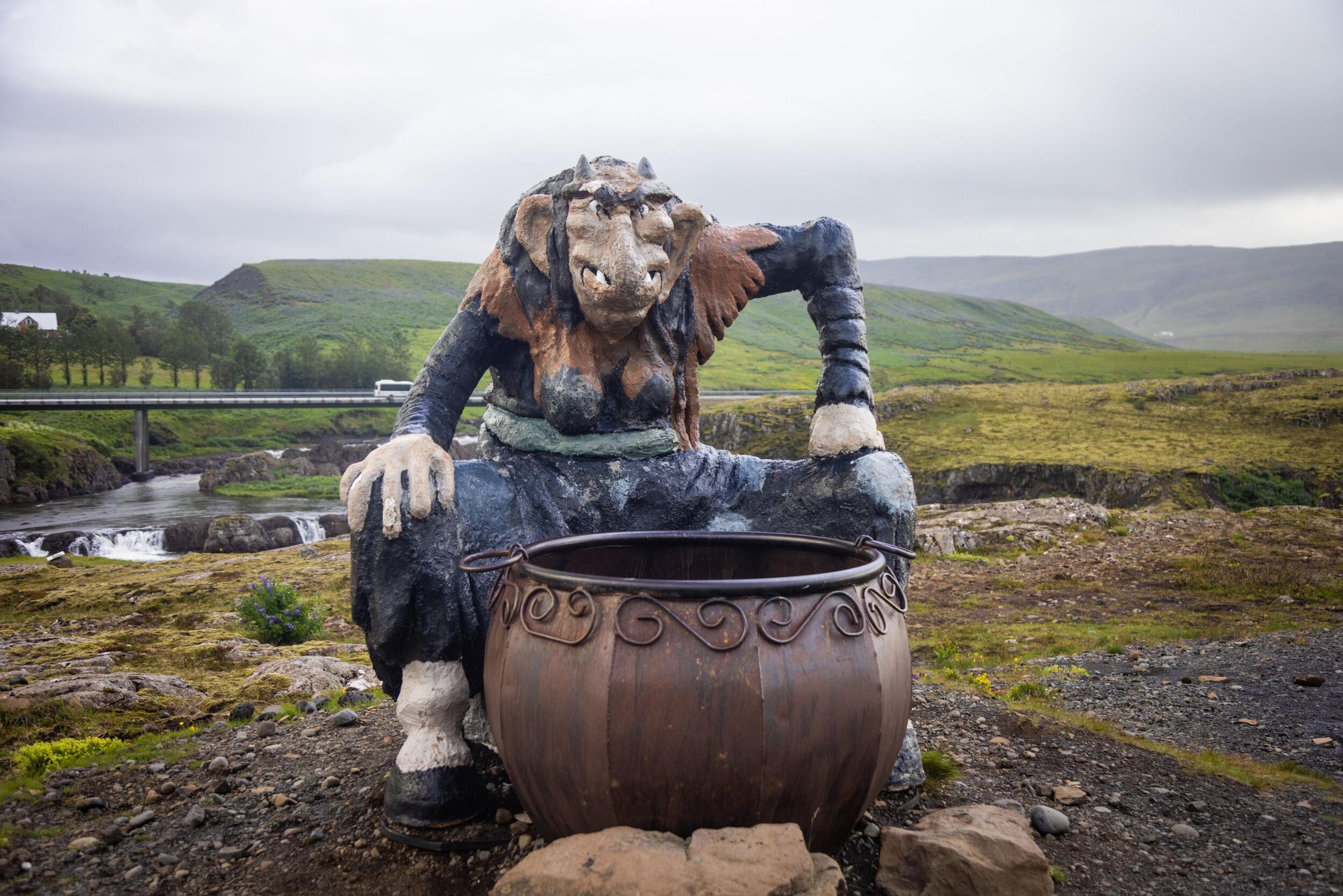 A large outdoor sculpture of a troll-like figure crouching over a cauldron, set against a lush Icelandic landscape with a bridge and waterfall in the background under a cloudy sky.