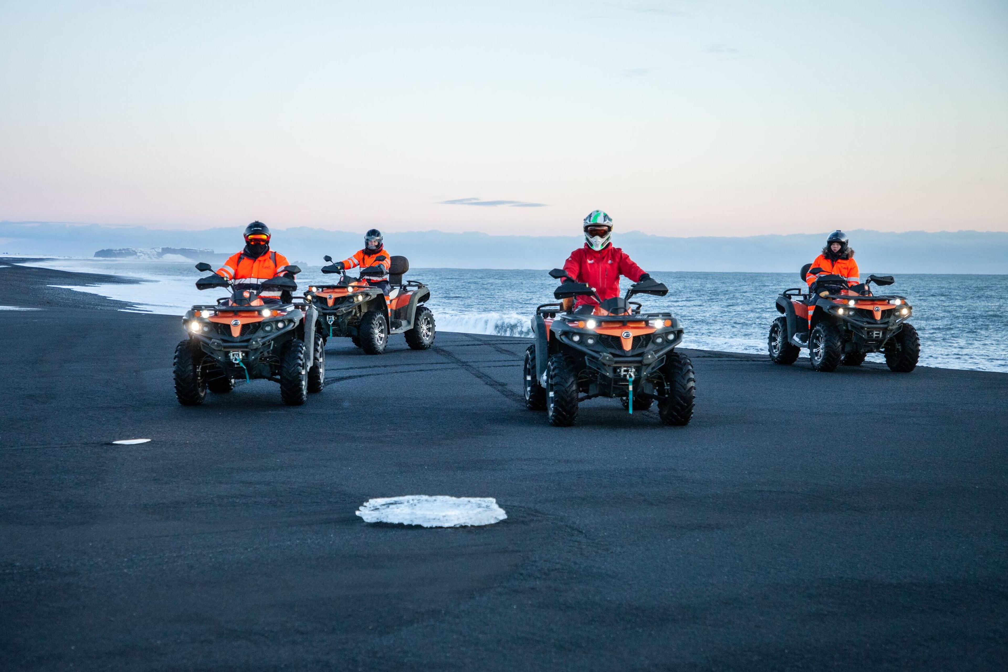 Four ATV riders in bright orange and red gear drive along a black sand beach near the ocean, with waves crashing beside them and a soft pastel sky in the background.