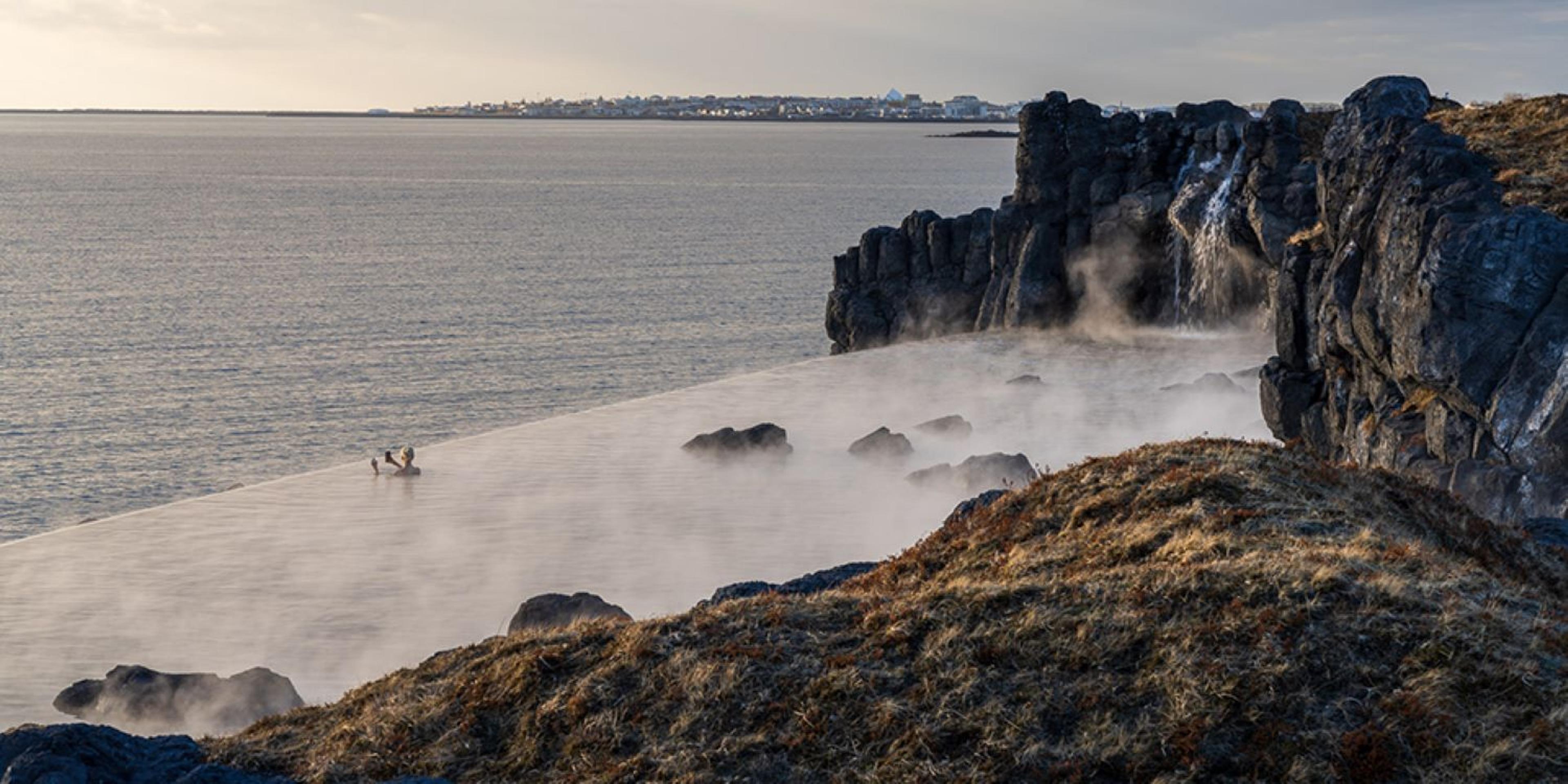 A steamy infinity pool with people, built into a rocky coastline with a waterfall, overlooking the ocean and a distant city skyline.
