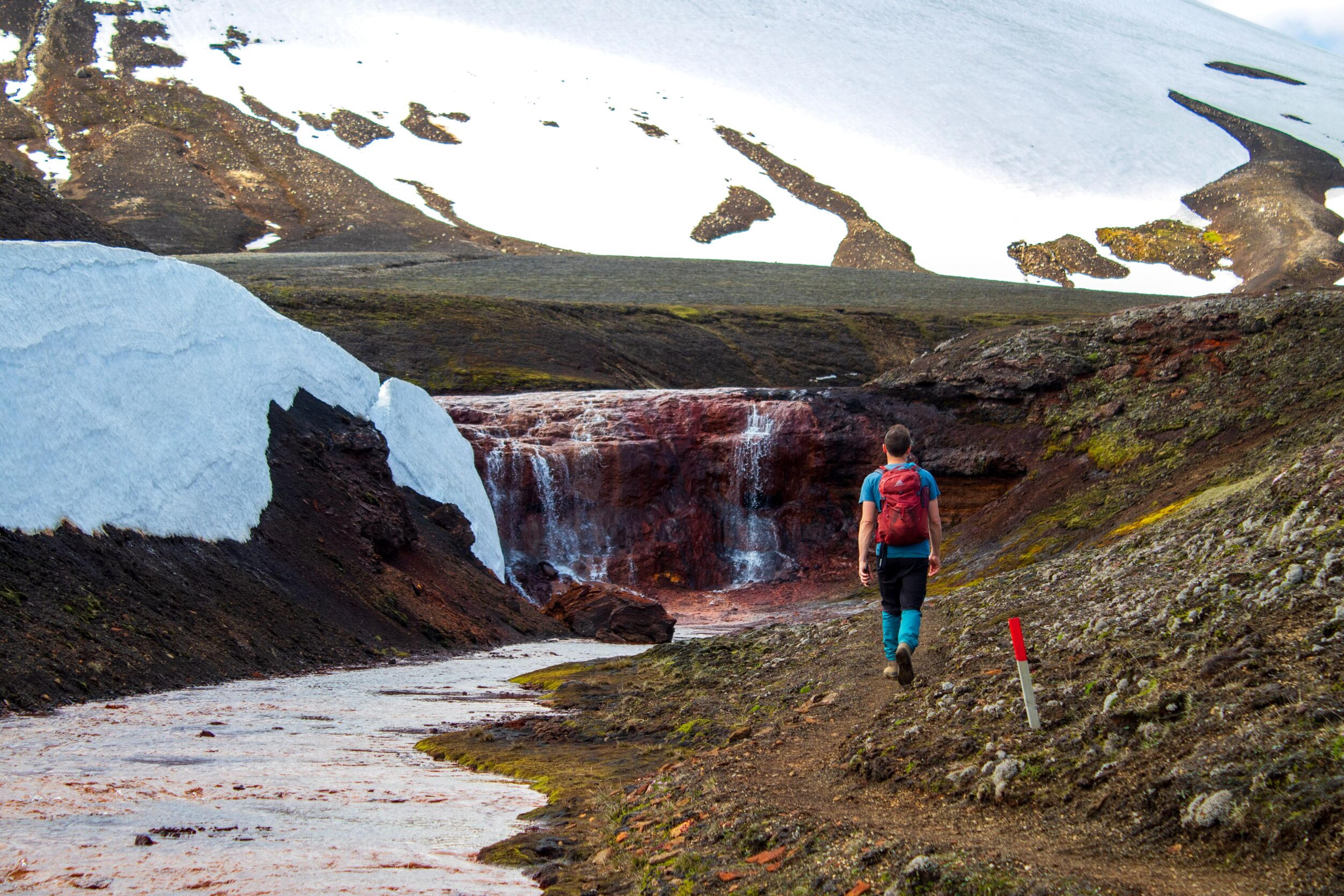 A person with a red backpack walks towards a reddish waterfall and stream in a snowy, rocky landscape.